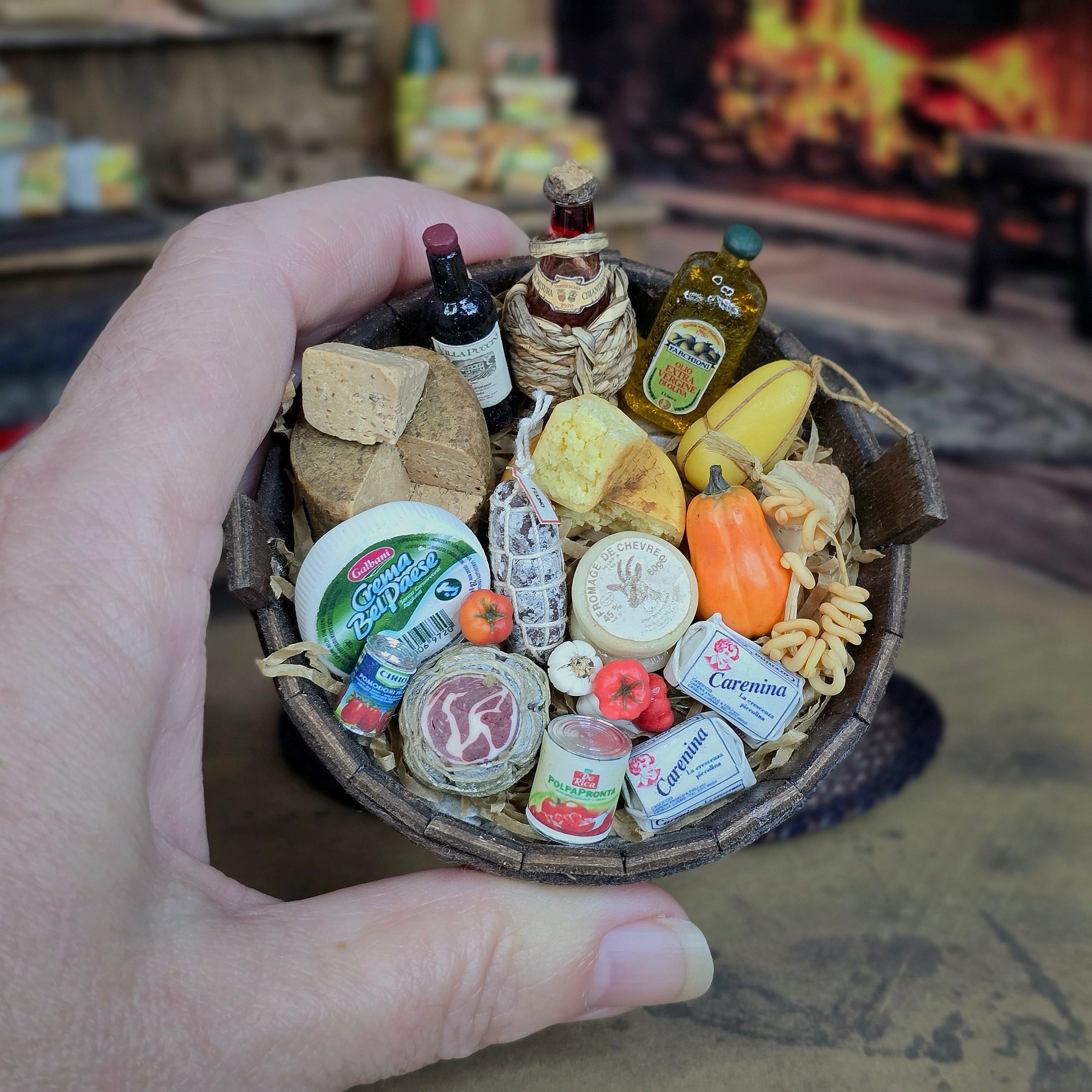 Hand holding a miniature basket filled with various food items and bottles against a blurred background.