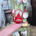 Load image into Gallery viewer, Red decorative teapot held between fingers with blurred background
