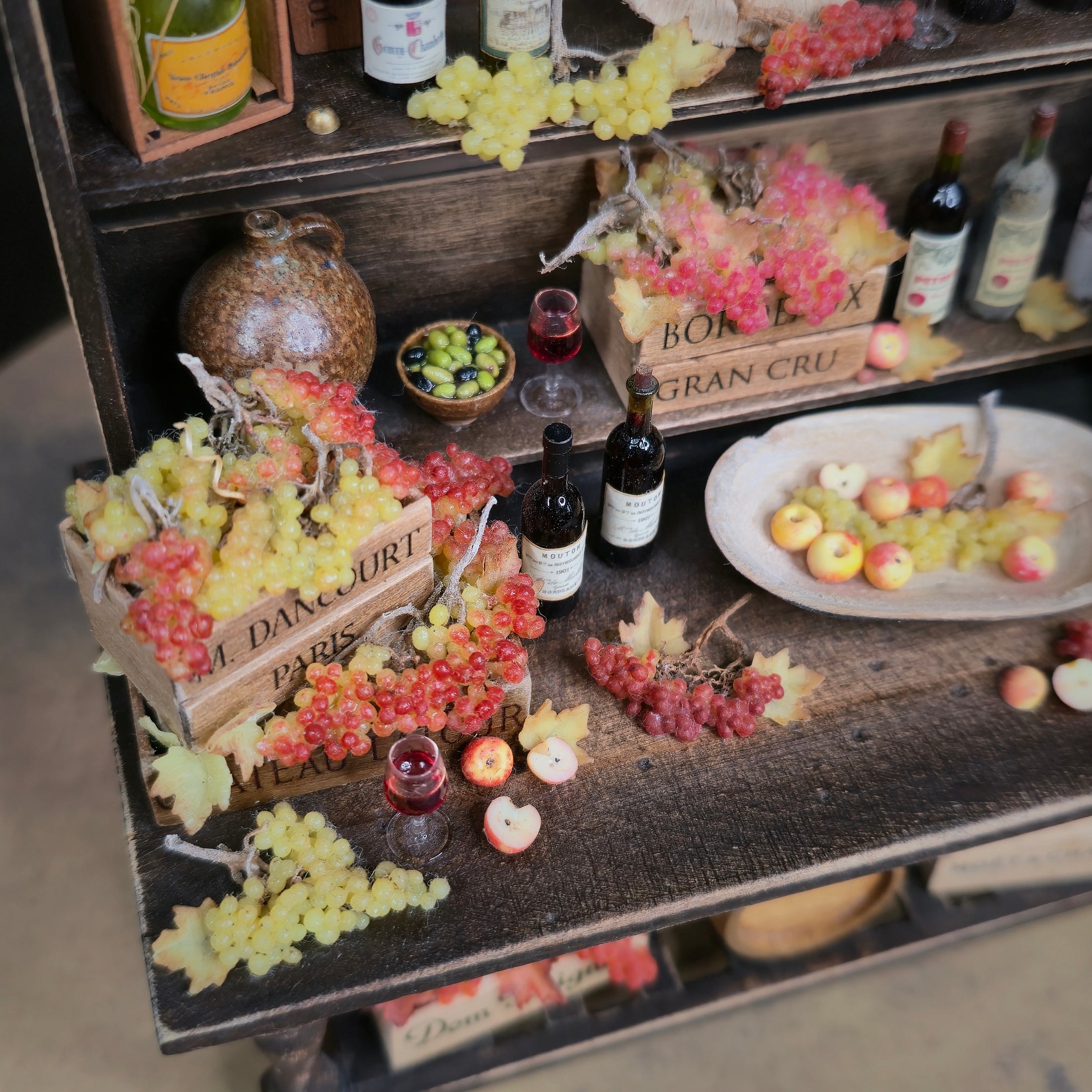Miniature table setup with fruits, bottles, and crates on a wooden surface.