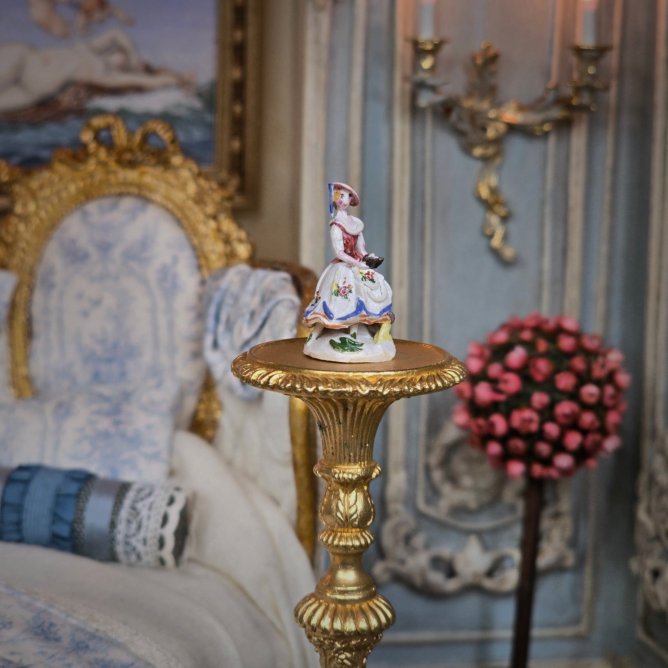 Decorative figurine on a gold pedestal in an ornate room with a chair and floral arrangement.
