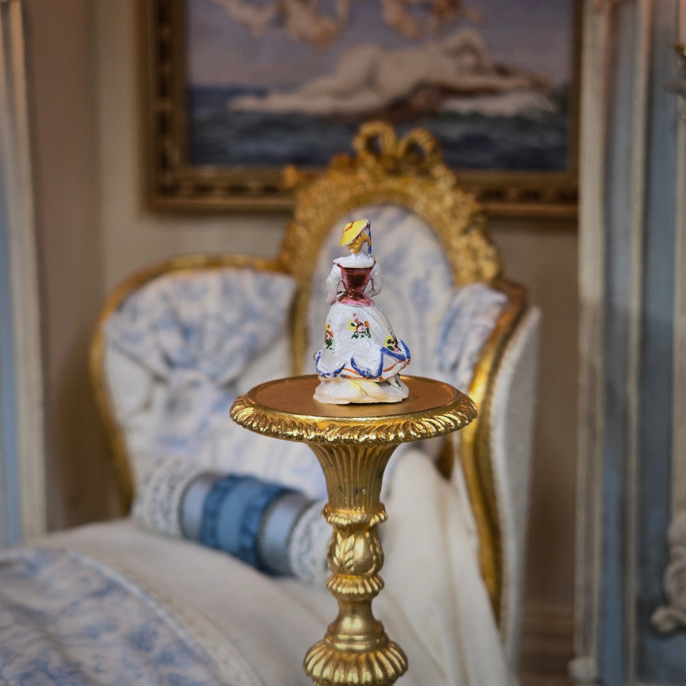 Decorative figurine on a gold pedestal table with a classical painting in the background