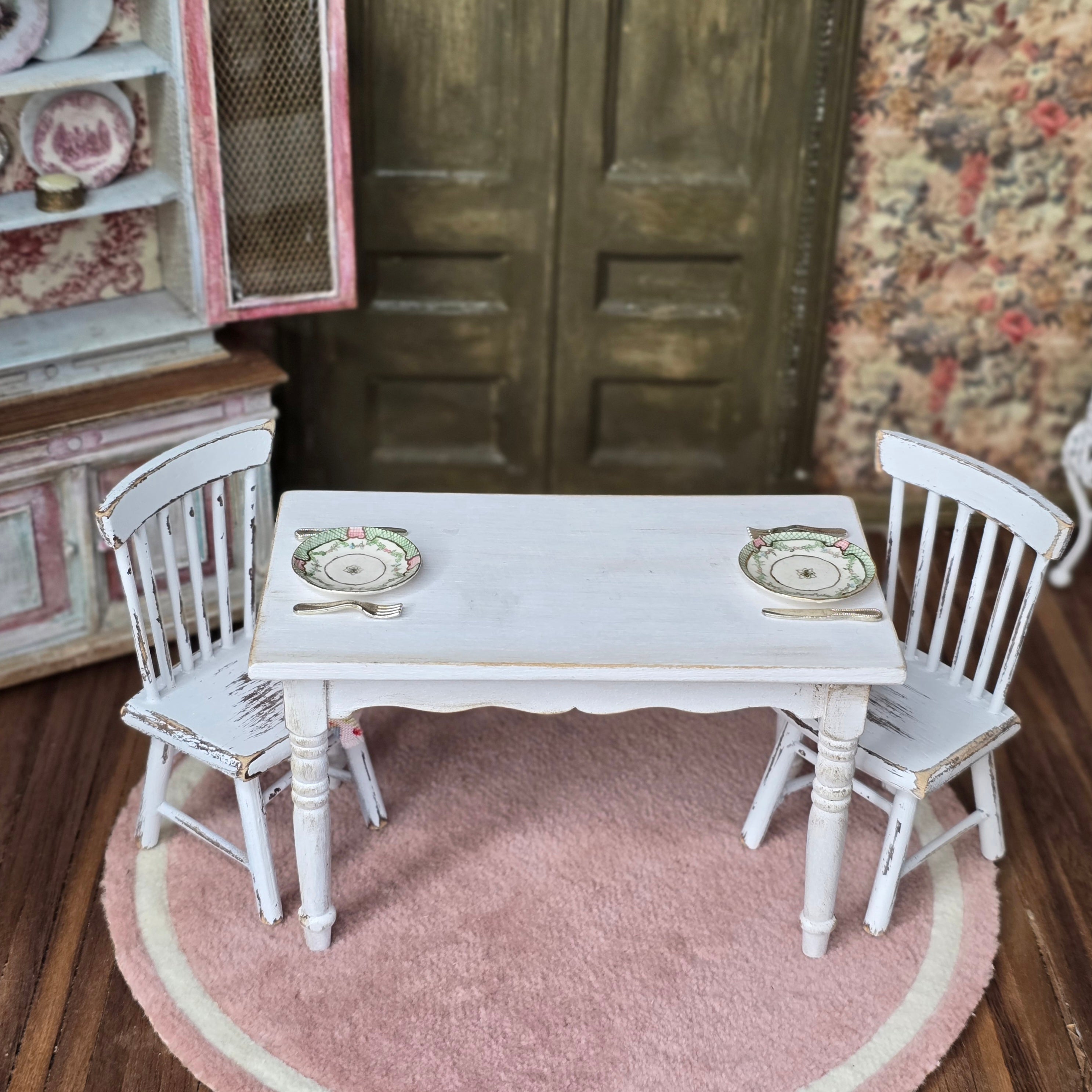 Miniature white table with two chairs on a pink rug in a dollhouse setting.