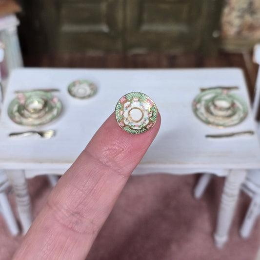 Close-up of a decorative SAUCER held by a finger with a miniature table and chairs in the background.