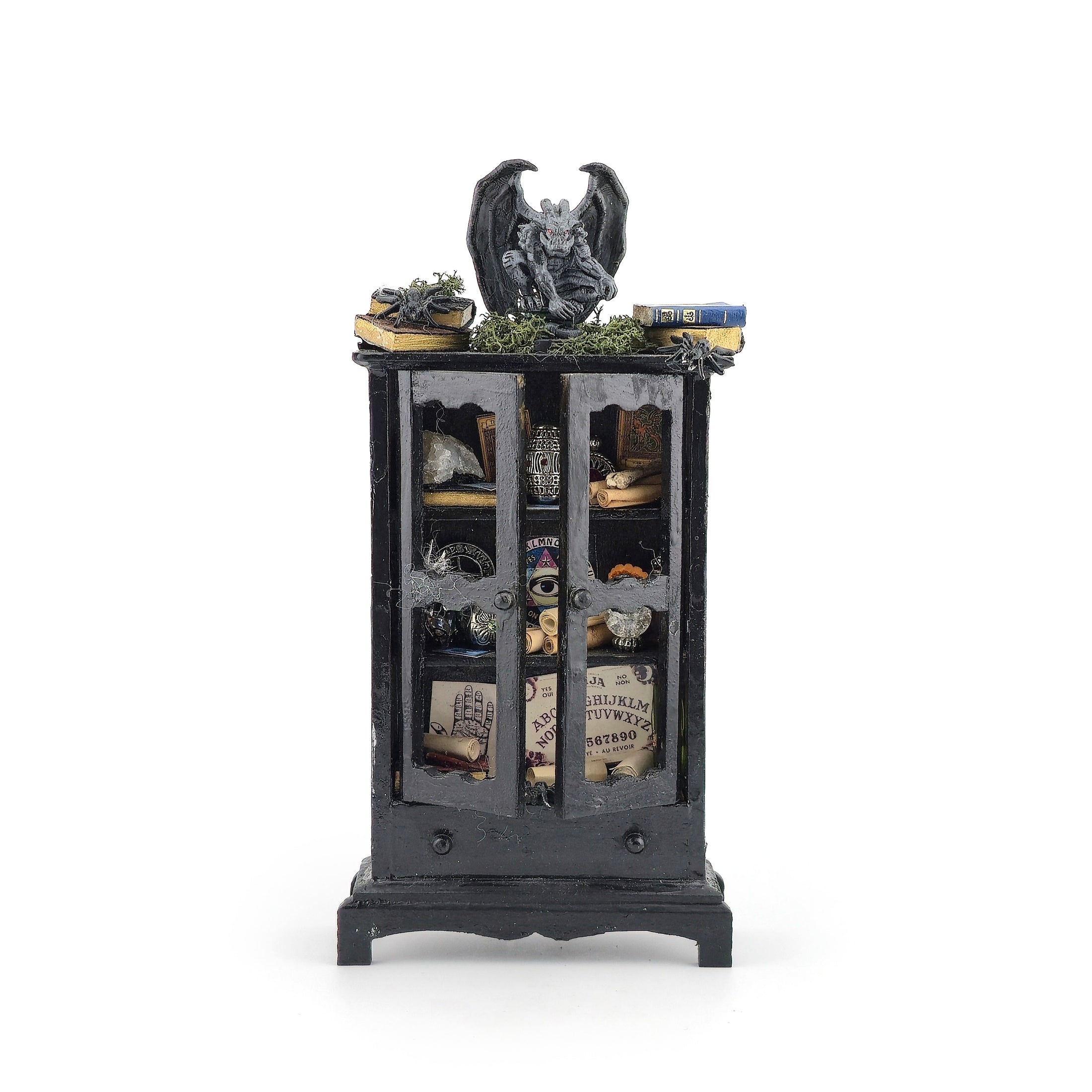 A miniature gothic-style black cabinet display with glass doors, filled with small decorative items including a gargoyle figurine, books, crystals, and old papers, shown on a plain white background.