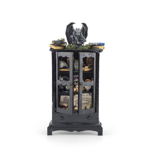A miniature gothic-style black cabinet display with glass doors, filled with small decorative items including a gargoyle figurine, books, crystals, and old papers, shown on a plain white background.