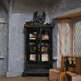 Load image into Gallery viewer, A tall black wooden display cabinet with doors stands in a rustic room with gray stone walls. The cabinet is styled with books, old papers, small figurines, and a dark gargoyle sculpture on top.
