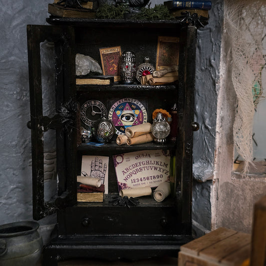A gothic-style black wooden cabinet with a glass door, filled with occult props including a Ouija board, crystal ball, tarot cards, an eye-of-providence card, and scrolls, set against a textured gray wall.