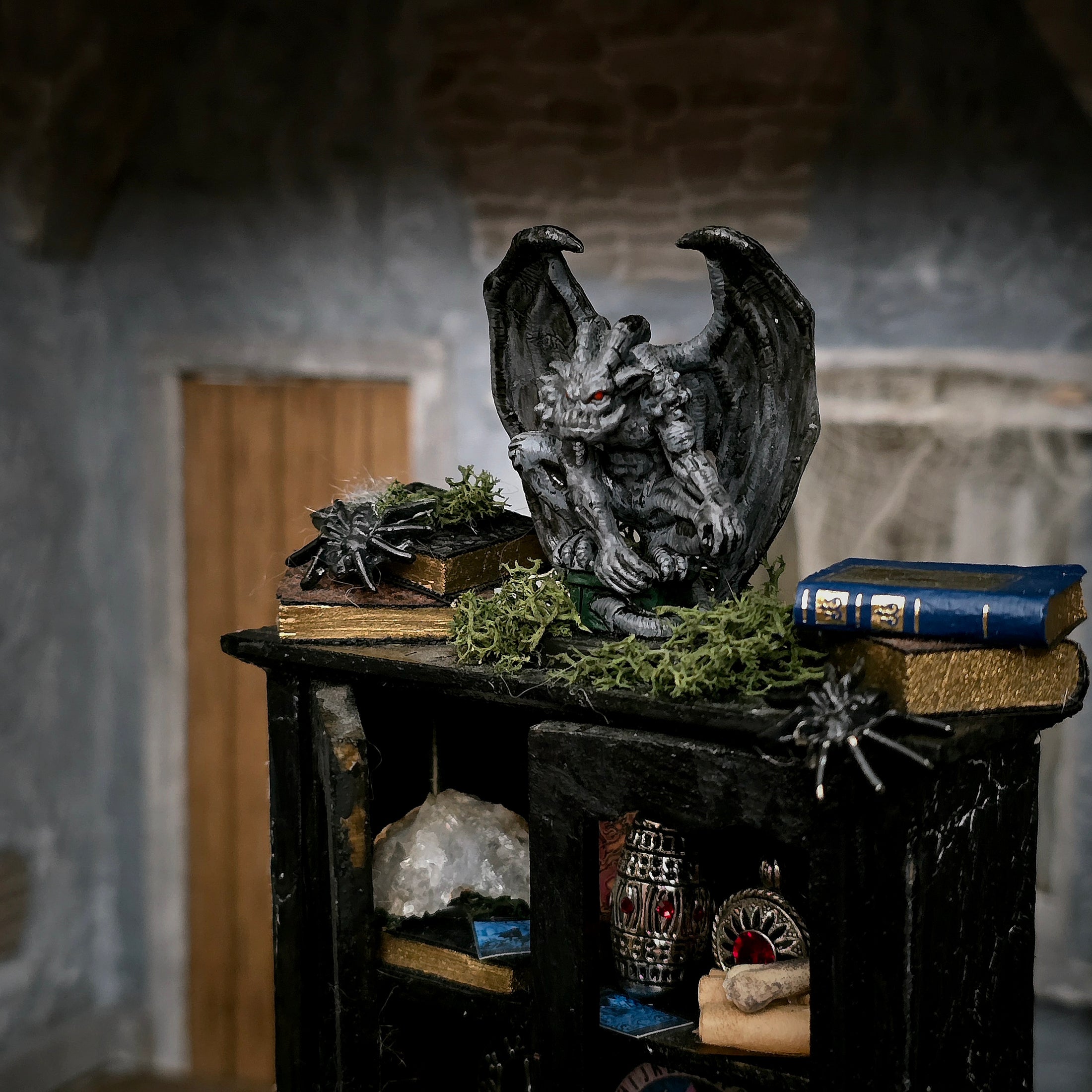 A gothic gargoyle figurine with outstretched wings and red eyes, displayed on a black shelf with old books, moss, and spider decorations in a stone room setting.