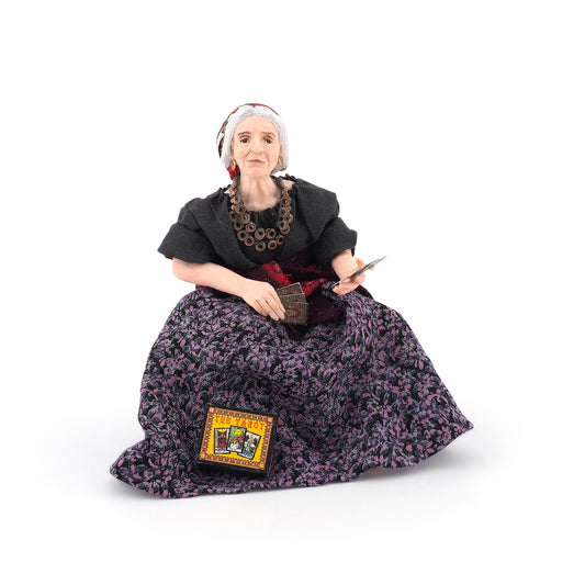 A clay dollhouse doll who is an elderly woman with gray hair, wearing a black top and a dark purple floral skirt, holding tarot cards, set against a plain white background.