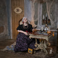 Load image into Gallery viewer, A woman in a black blouse and a dark purple floral skirt sits in a gothic, mystical room with a wooden table, tarot cards, candles, and cobweb decorations.
