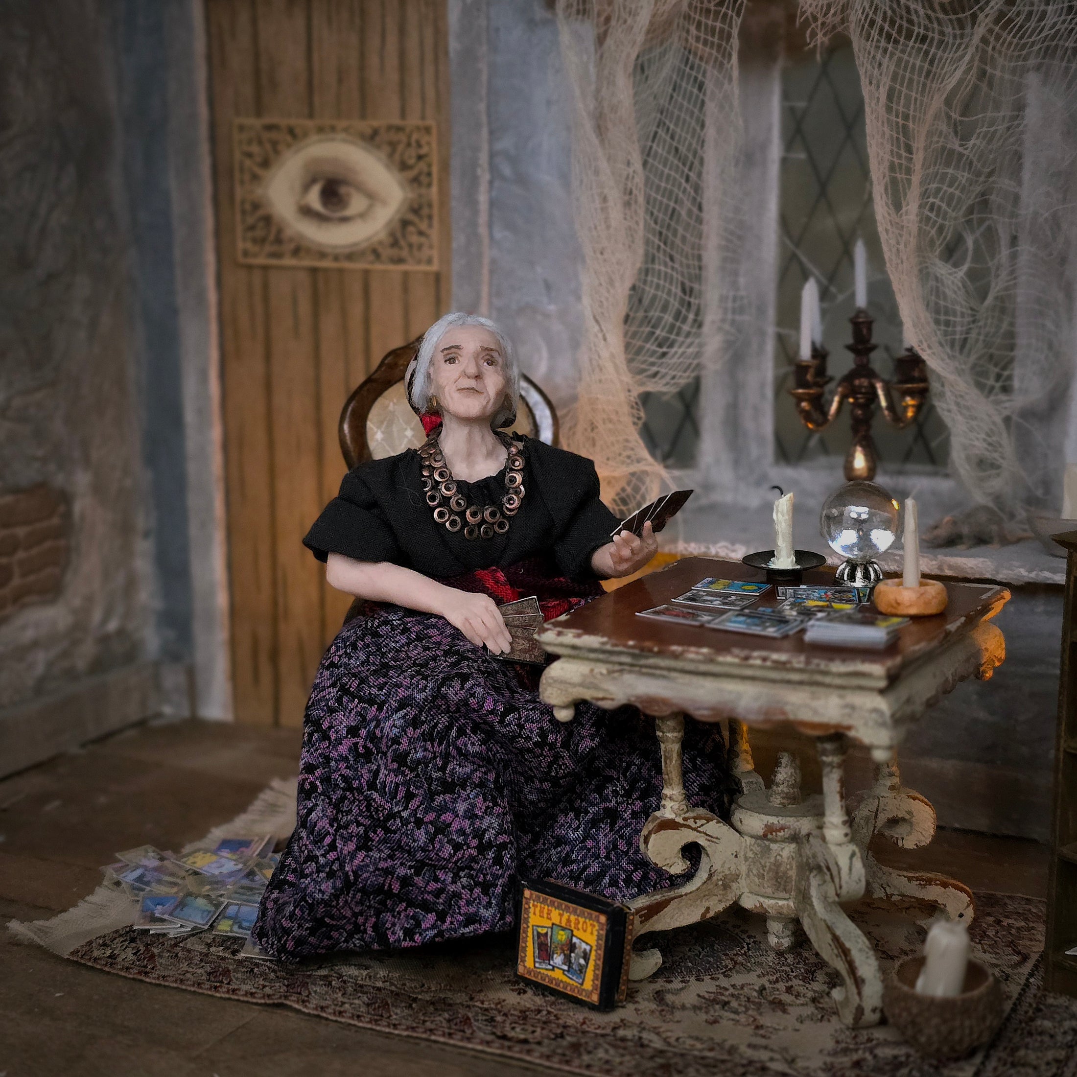 A woman in a black blouse and a dark purple floral skirt sits in a gothic, mystical room with a wooden table, tarot cards, candles, and cobweb decorations.