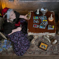 Load image into Gallery viewer, An elderly woman dollhouse doll with gray hair and a red headscarf sits at a small wooden table surrounded by tarot cards, candles, and ritual objects in a dimly lit room.

