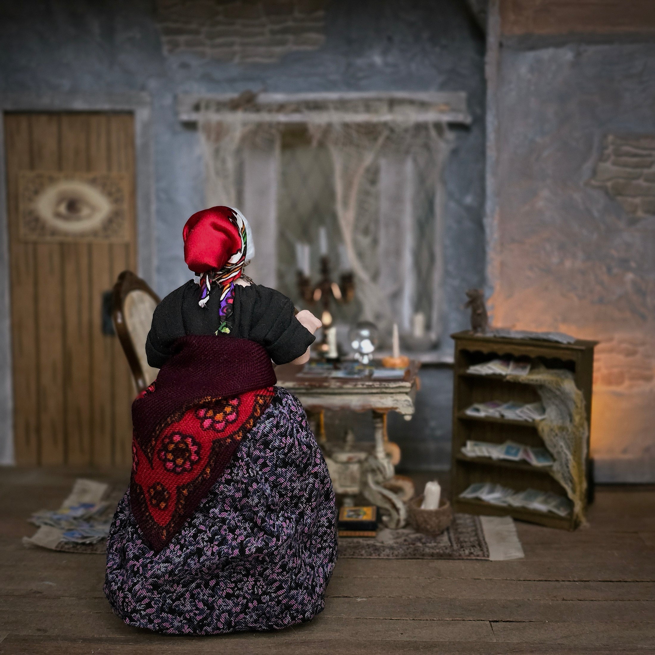The backside view or a gypsy woman tarot card character dollhouse doll in a vintage-style costume with a red headscarf, black top, and a purple floral skirt with a red patterned sash, seated in a dimly lit, rustic room with a table, candles, and shelves.