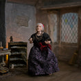 Load image into Gallery viewer, An elderly woman dollhouse doll with white hair wearing a black top, a purple floral skirt, and a red head covering stands in a rustic, dimly lit room with wooden floors and old furniture.
