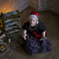 Load image into Gallery viewer, An elderly woman with gray hair, wearing a red headscarf, black top, and a purple patterned skirt, sitting on a wooden floor in a dimly lit room and holding tarot cards.
