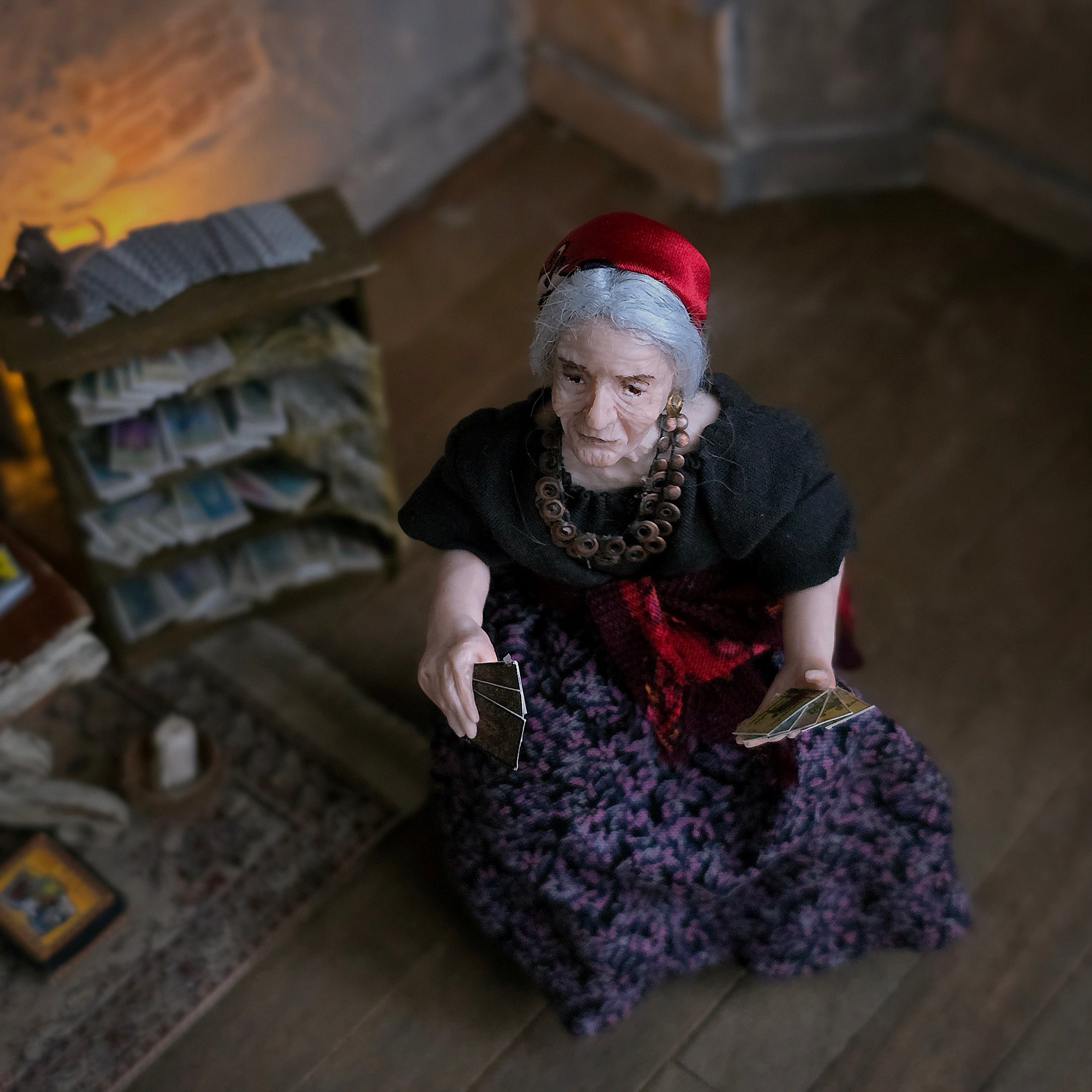 An elderly woman with gray hair, wearing a red headscarf, black top, and a purple patterned skirt, sitting on a wooden floor in a dimly lit room and holding tarot cards.