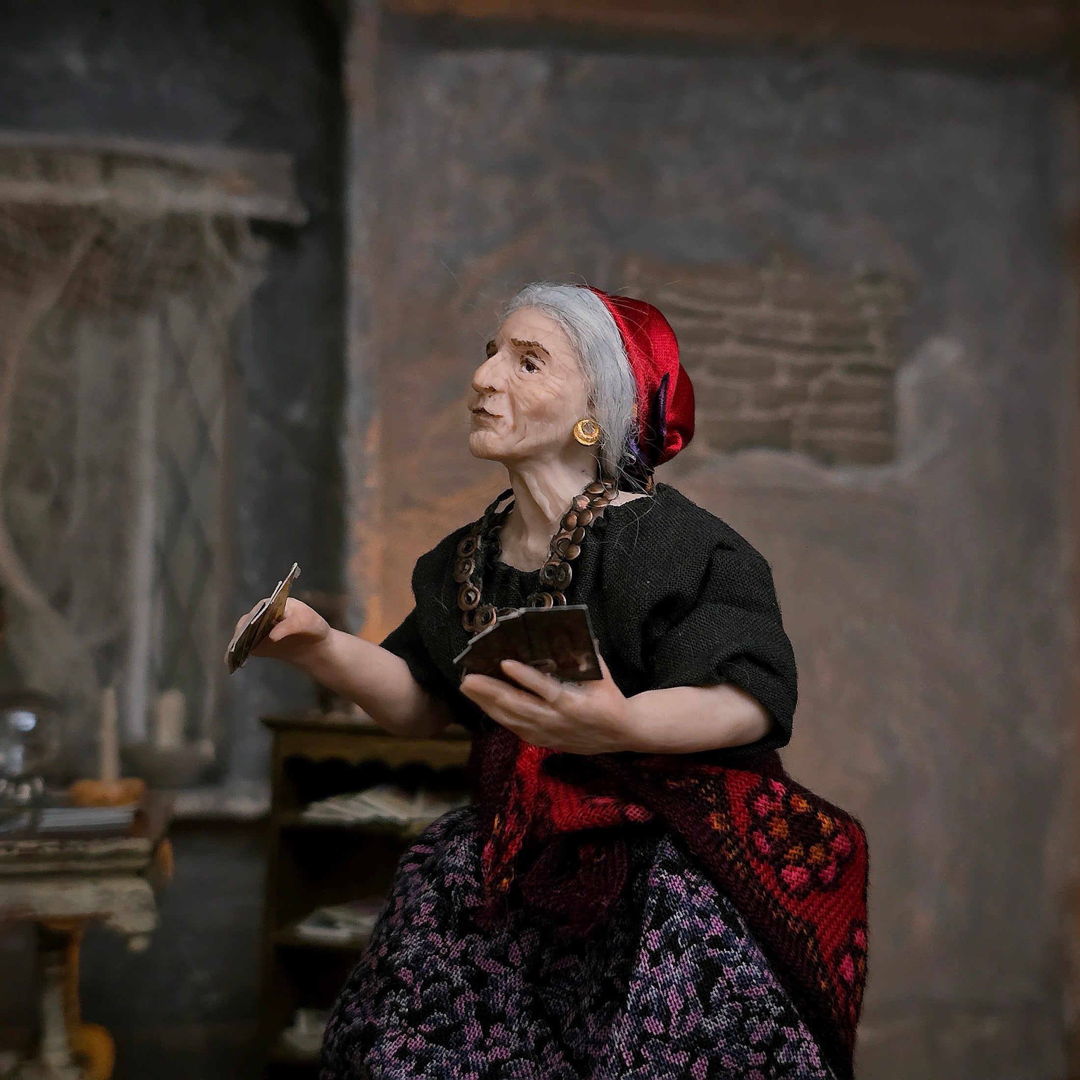 A detailed miniature elderly woman doll with gray hair, wearing a red headscarf, black top, and a purple patterned skirt, holding tarot cards in a dimly lit room.