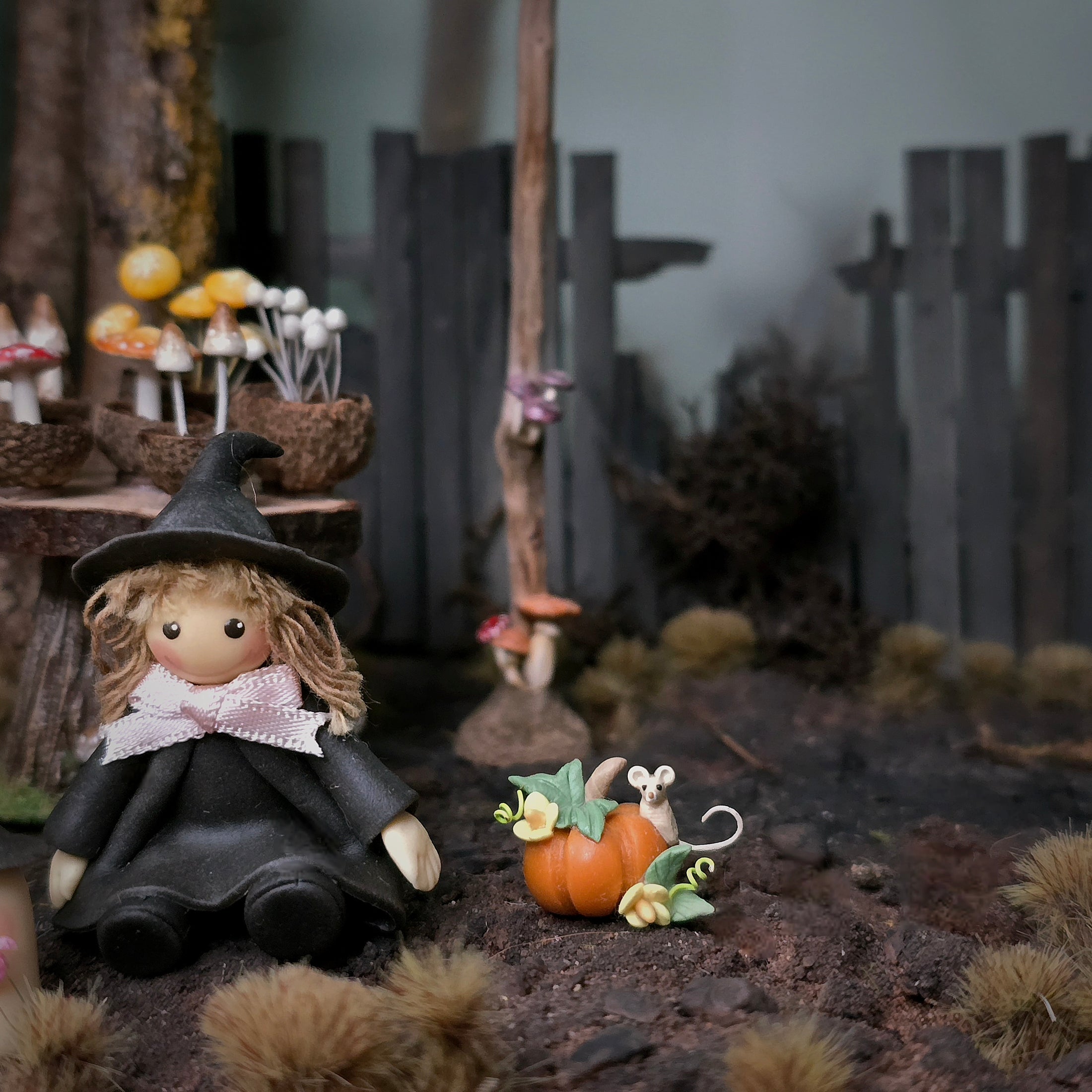 A whimsical miniature fairy garden scene featuring a witch doll in a black hat and dress sitting beside a small orange pumpkin with a tiny mouse, set against a rustic wooden fence and woodland backdrop.