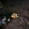 Load image into Gallery viewer, A miniature orange pumpkin ornament decorated with green leaves, small yellow flowers, and a tiny white rabbit figure, placed on dark textured soil next to a small figurine.
