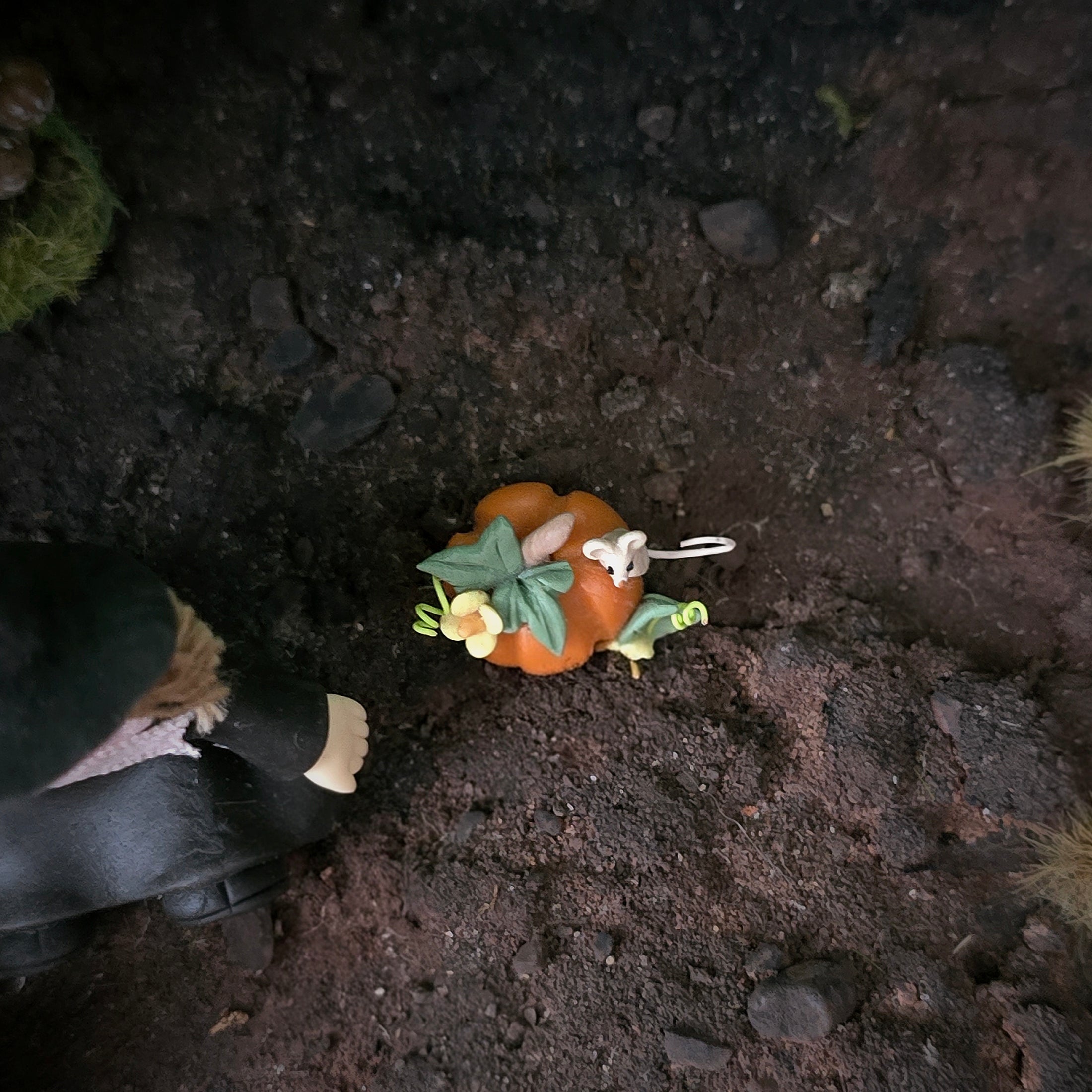 A miniature orange pumpkin ornament decorated with green leaves, small yellow flowers, and a tiny white rabbit figure, placed on dark textured soil next to a small figurine.