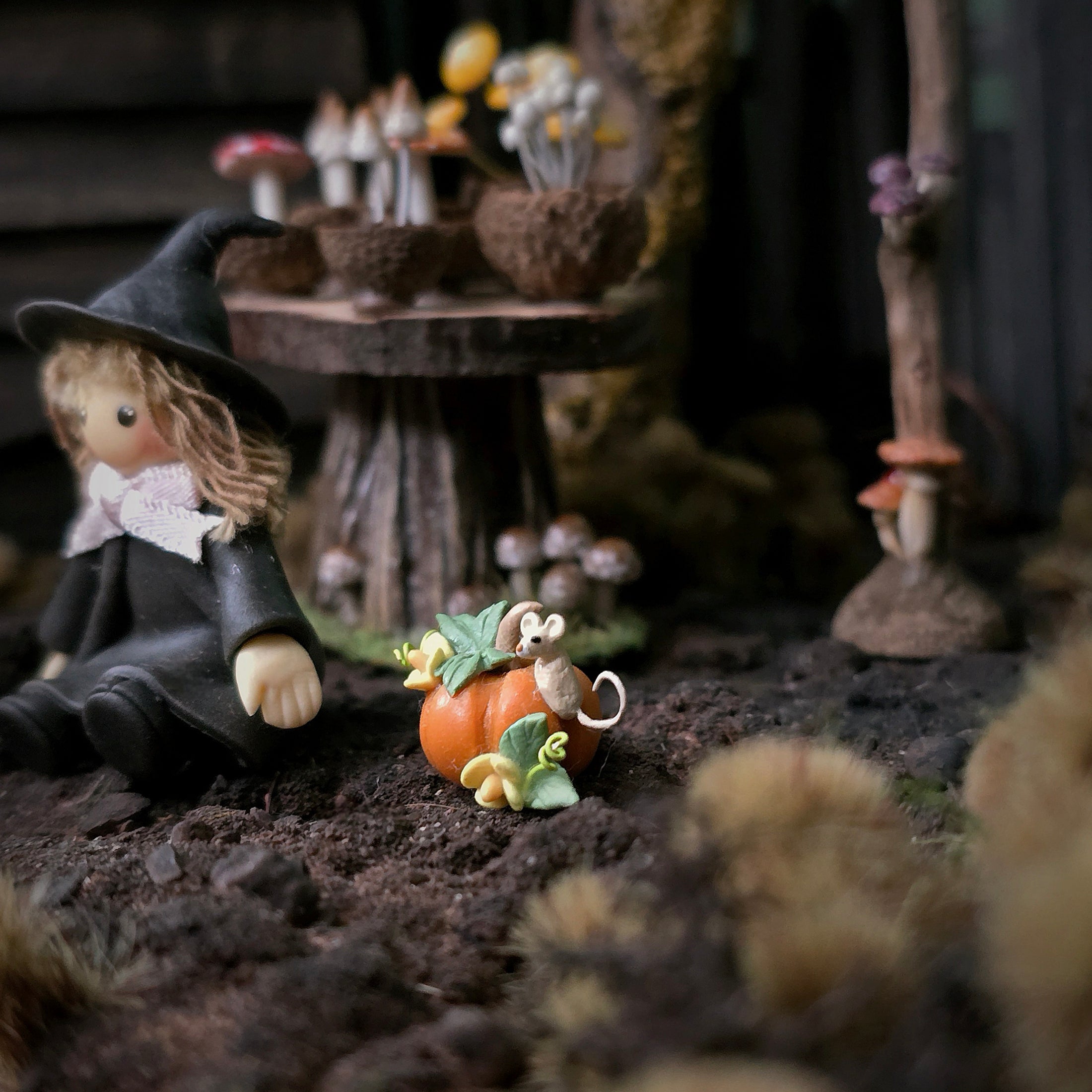A miniature fairy garden scene with a small witch doll in a black dress and hat beside a tiny orange pumpkin with green leaves, set in a dark forest-like diorama with mushrooms and a wooden table in the background.