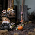 Load image into Gallery viewer, A miniature fairy garden scene with a small witch doll in a black hat and dress beside an orange pumpkin, with tiny mushrooms, a wooden fence, and dried grass in the background.
