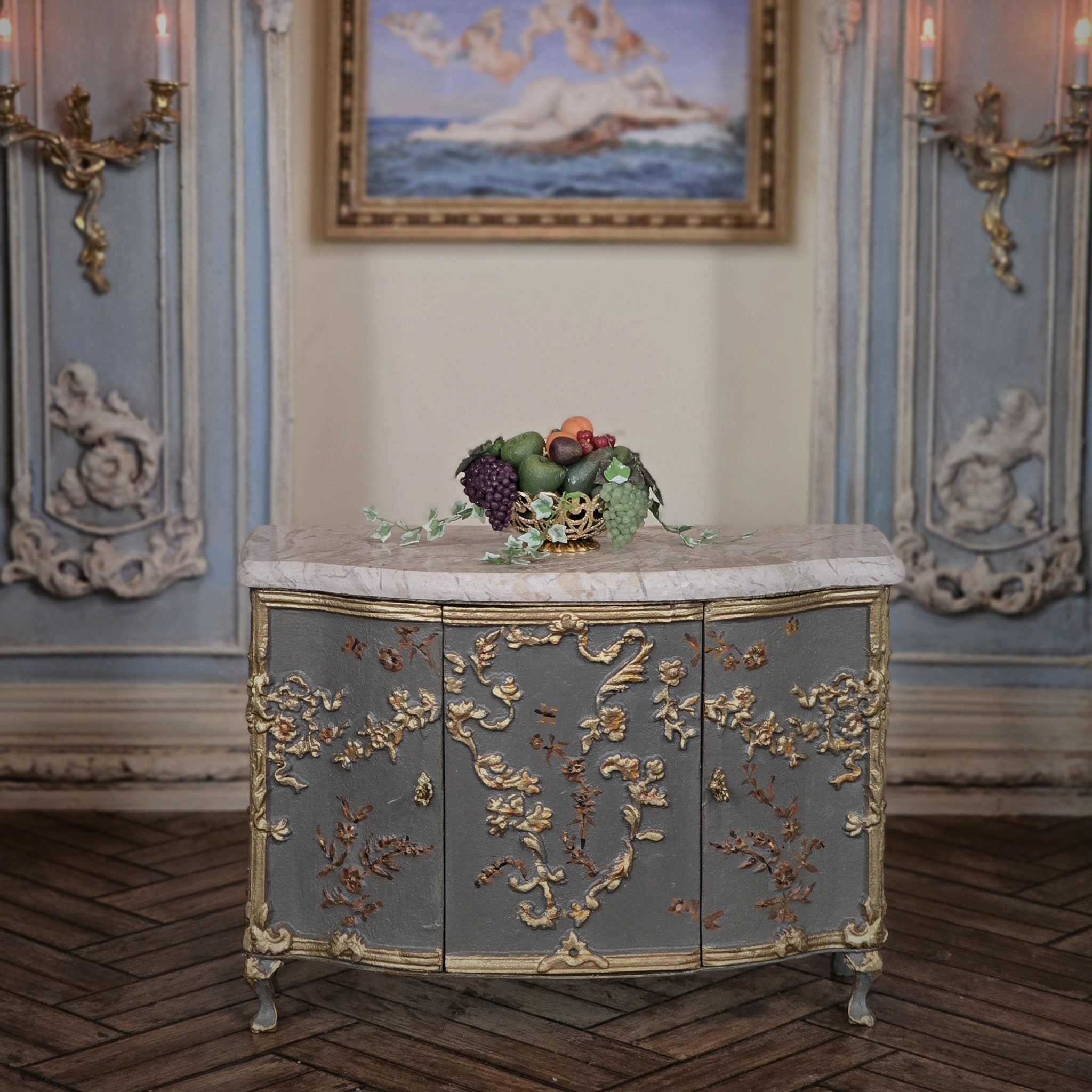 Decorative miniature dollhouse bowl of fruit on a cabinet with marble top and gold embellishments in an ornate room.