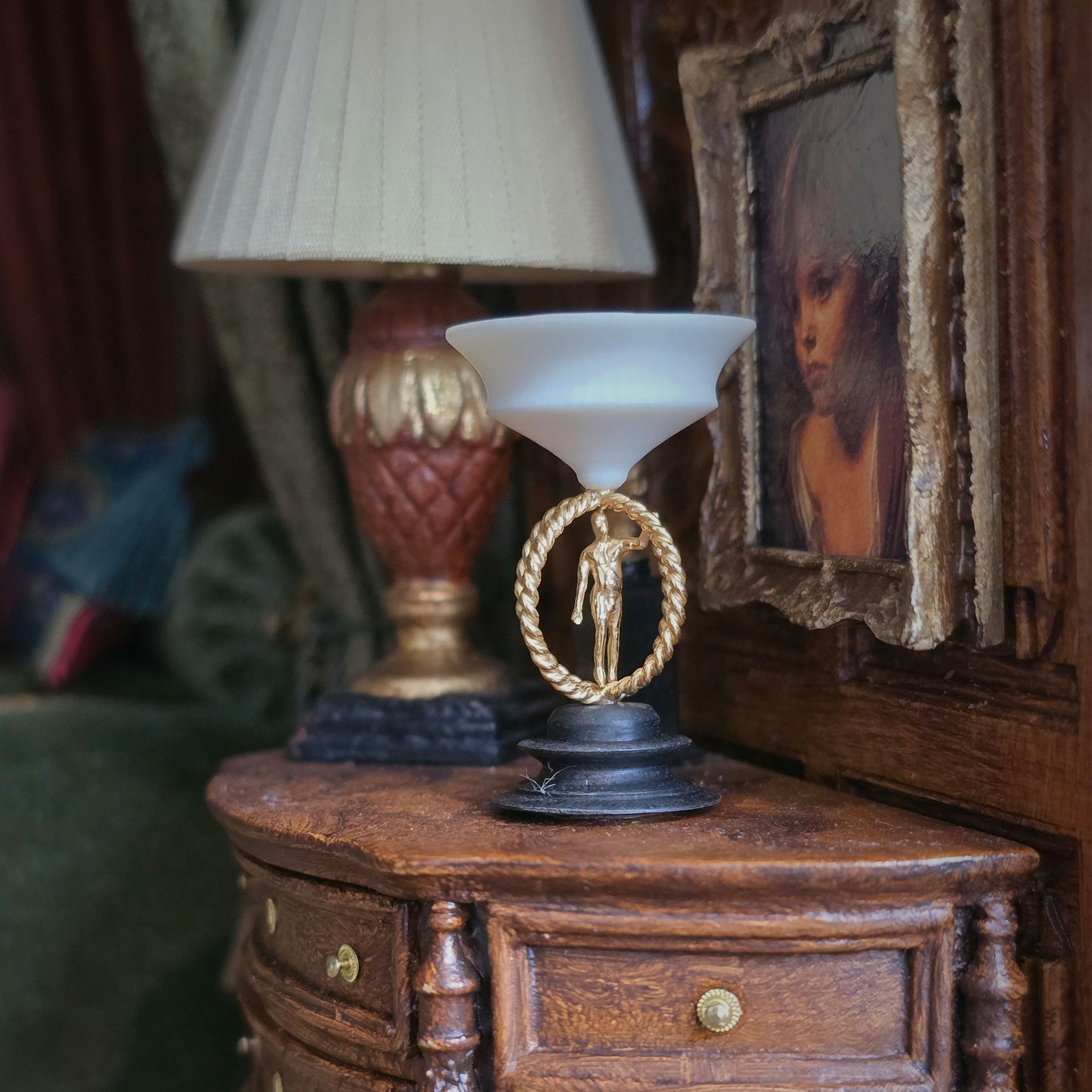 Decorative sculpture with a metal male figure on a black base in a warm, indoor miniature setting, with a lamp and framed picture in the background.