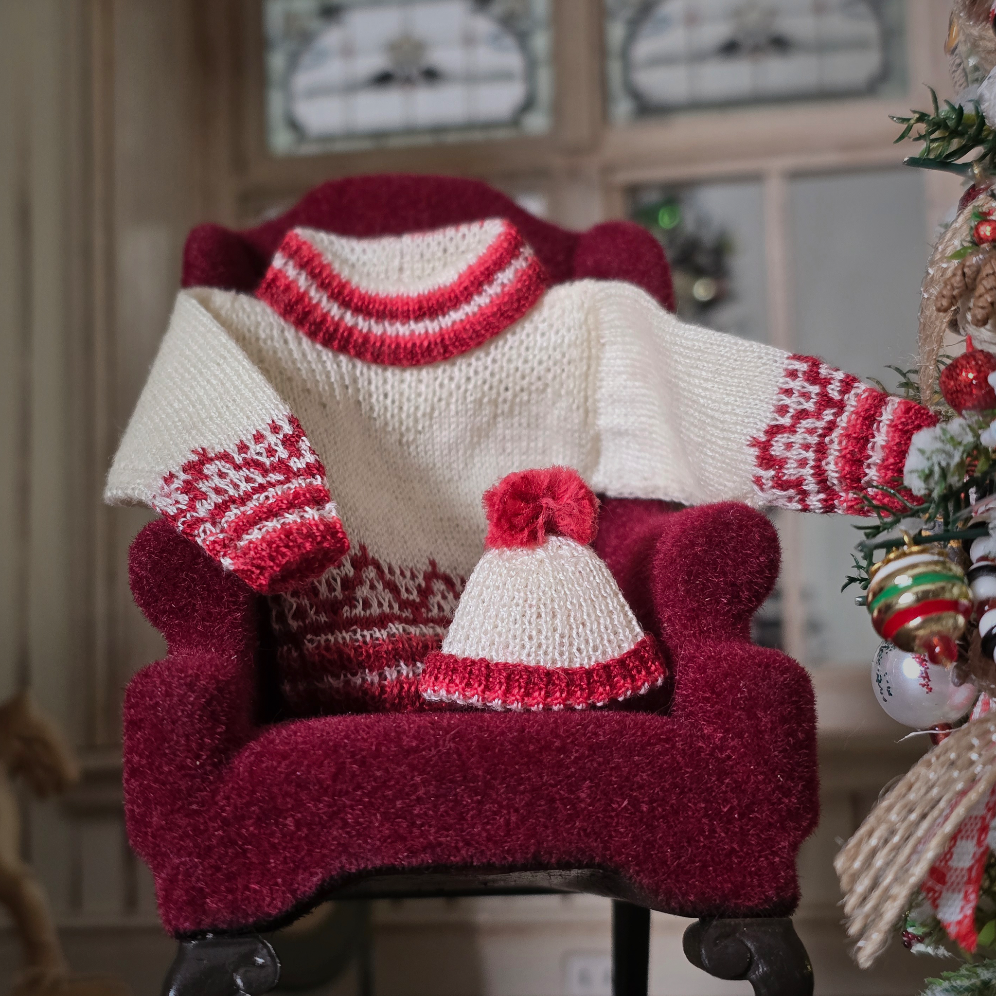 Dollhouse miniature Red and cream knitted sweater with matching hat on a red chair, Christmas tree in the background