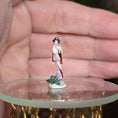 Load image into Gallery viewer, Small figurine of a woman in traditional attire on a round surface held in a hand.
