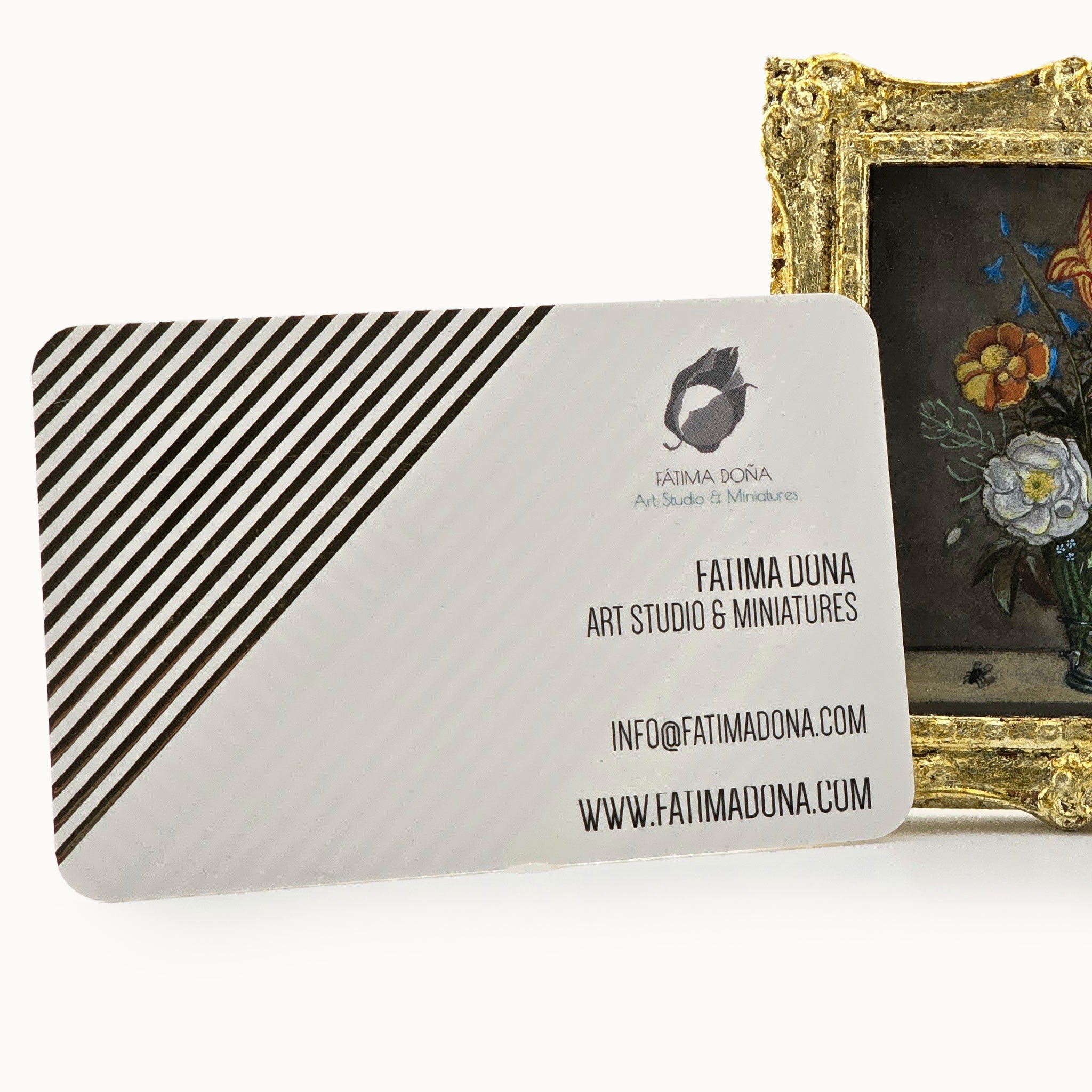 Business card with striped design and contact information for Fatima Dona Art Studio & Miniatures, placed next to a decorative gold frame with floral artwork.