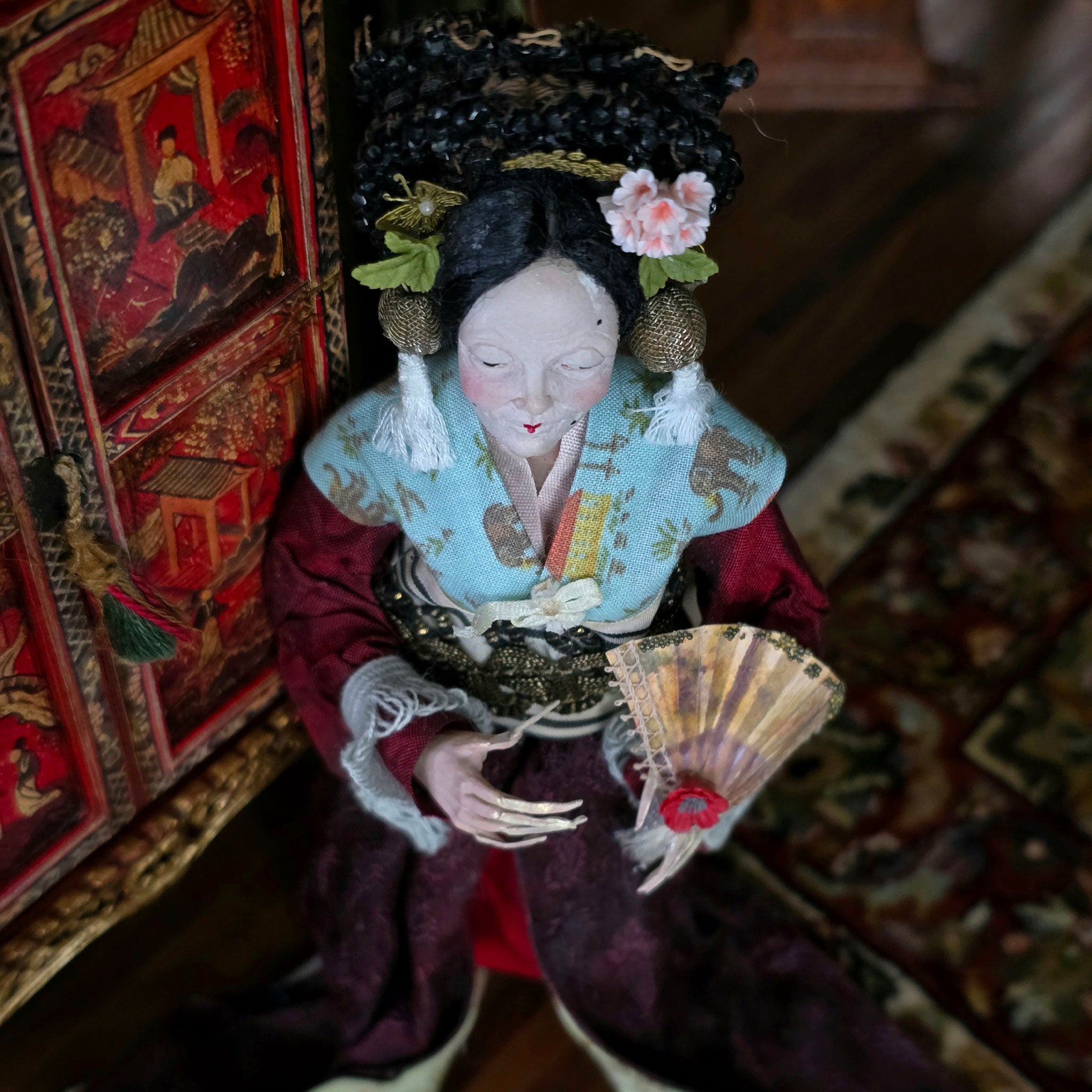 Traditional doll in ornate clothing holding a fan, with a blurred background