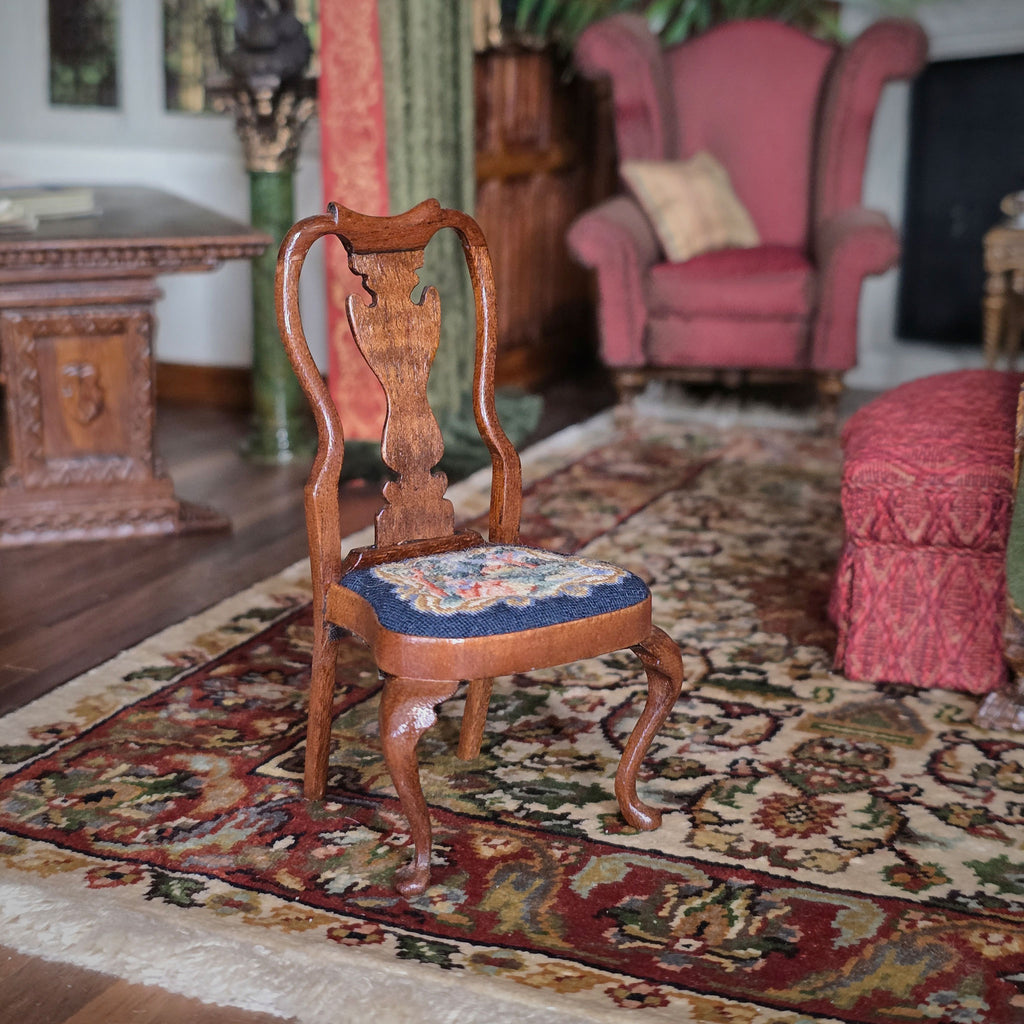 Wooden chair with a scenic needlepoint cushion on a patterned rug in a room with furniture and decor.
