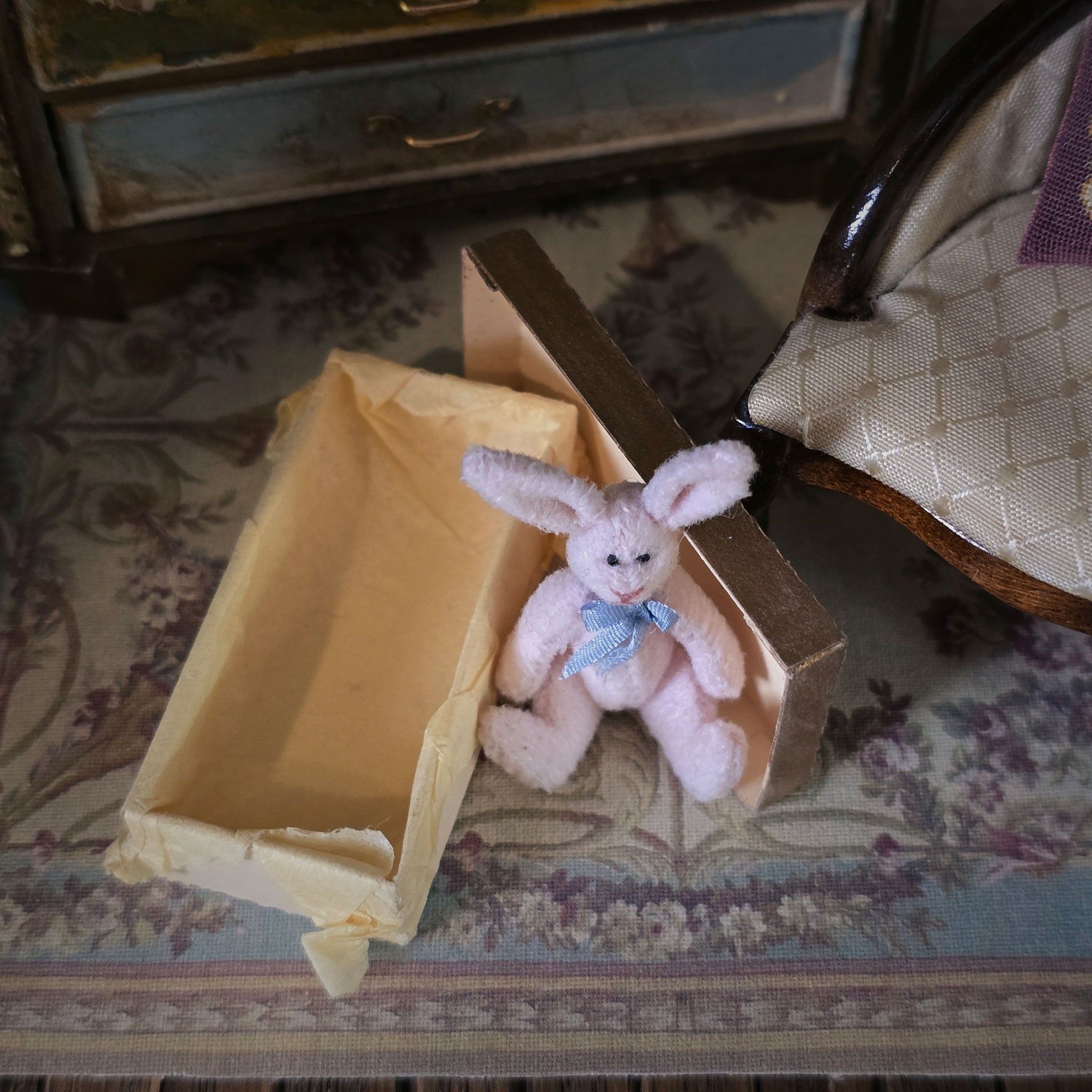 Small pink rabbit toy with a blue bow tie inside an open aged box on a patterned rug.