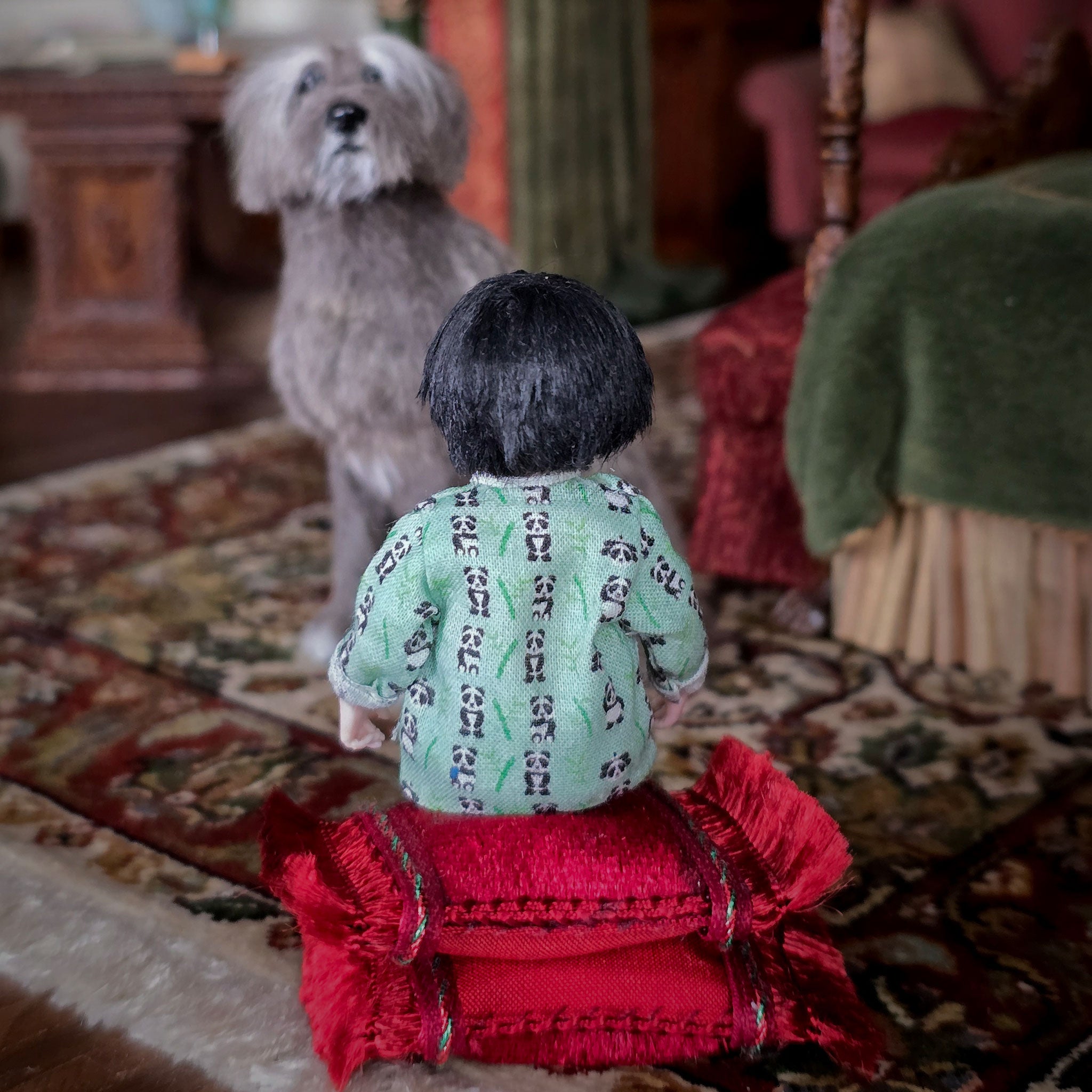 Small doll with a green top sitting on a red pillow with a dog in the background.