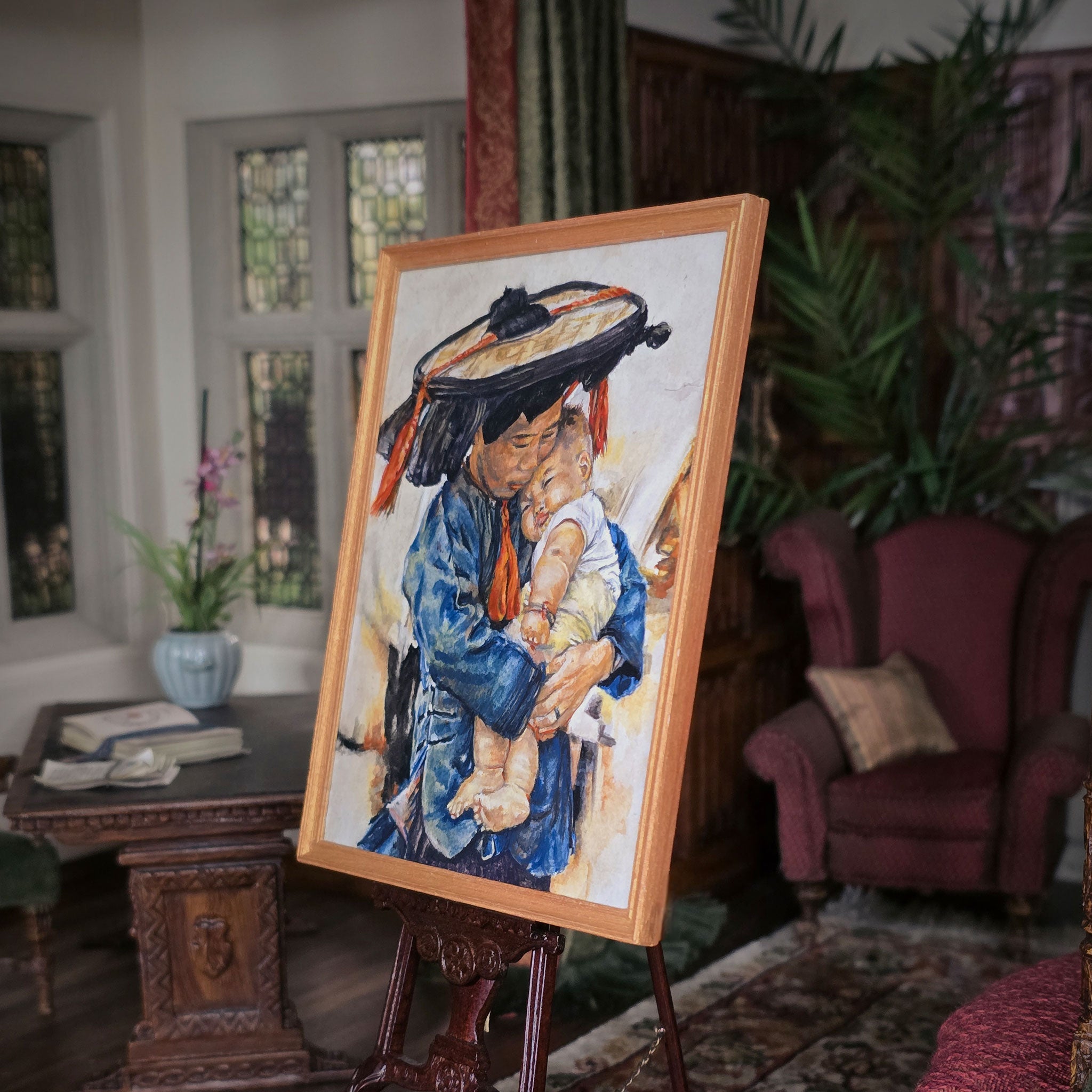 Painting of a woman holding a child on an easel in a room with furniture and plants. Deanna Karras