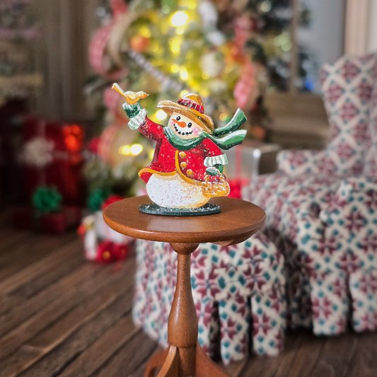 Decorative snowman figurine on a small wooden table with a blurred Christmas tree and chair in the background.