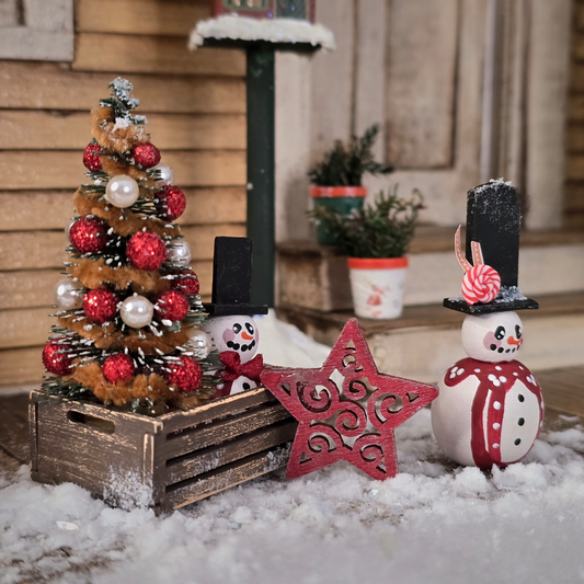 Small Christmas tree with red ornaments, snowmen, and a star decoration on a snowy surface.