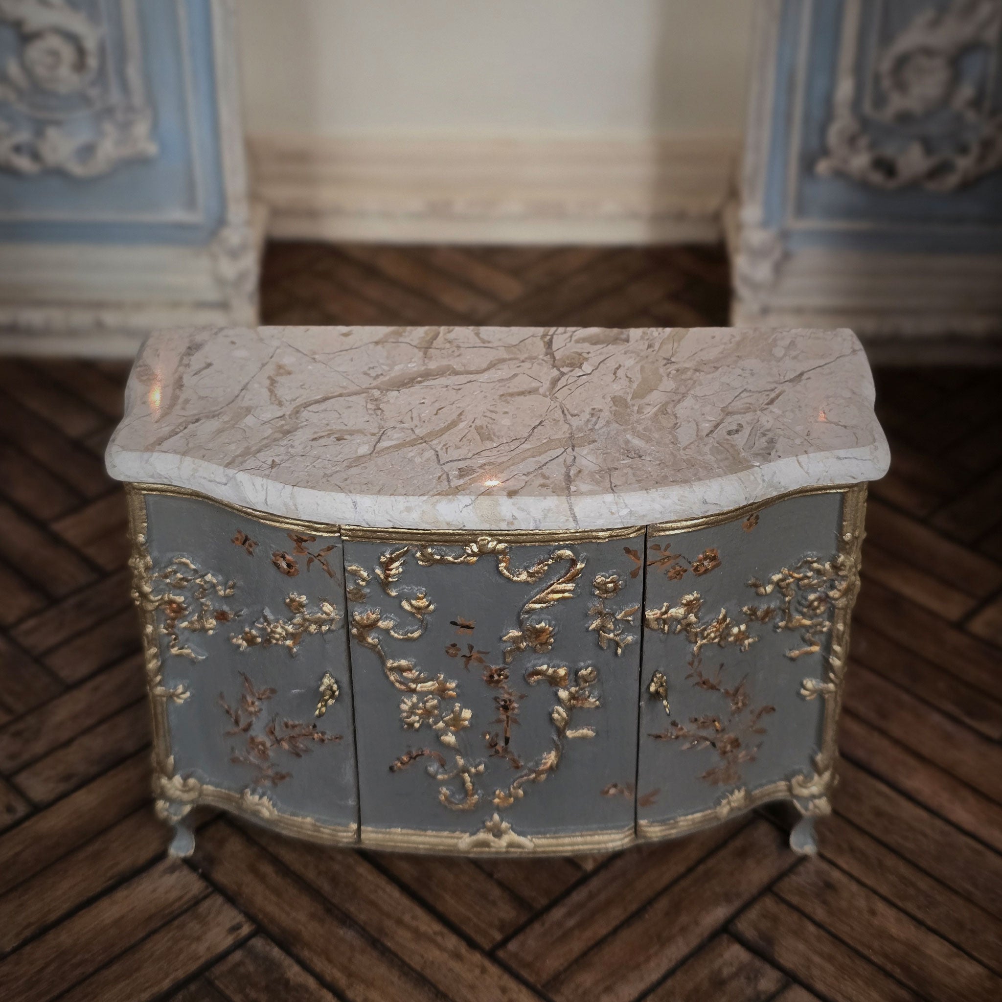 Decorative sideboard cabinet in miniature with marble top and gold leaf detailing on a wooden floor.