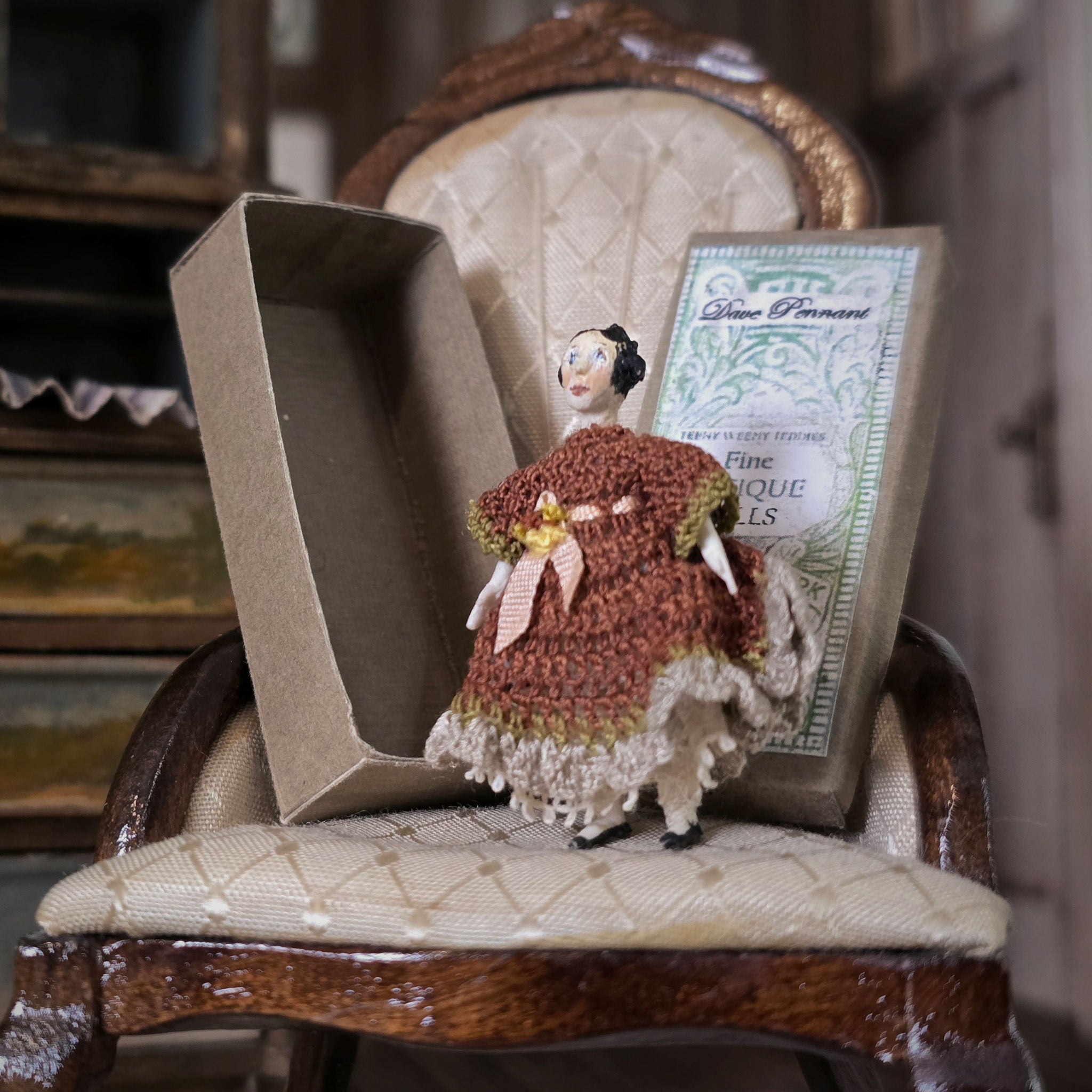 Small doll in a brown dress sitting on a vintage chair with a box in the background