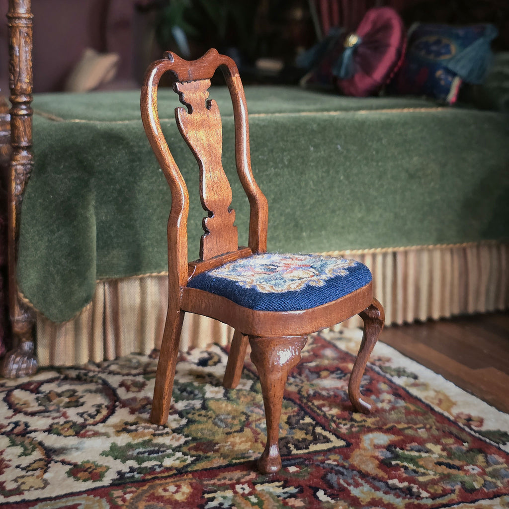Wooden chair with a petit point cushion on a rug in a room with a green bed in background.