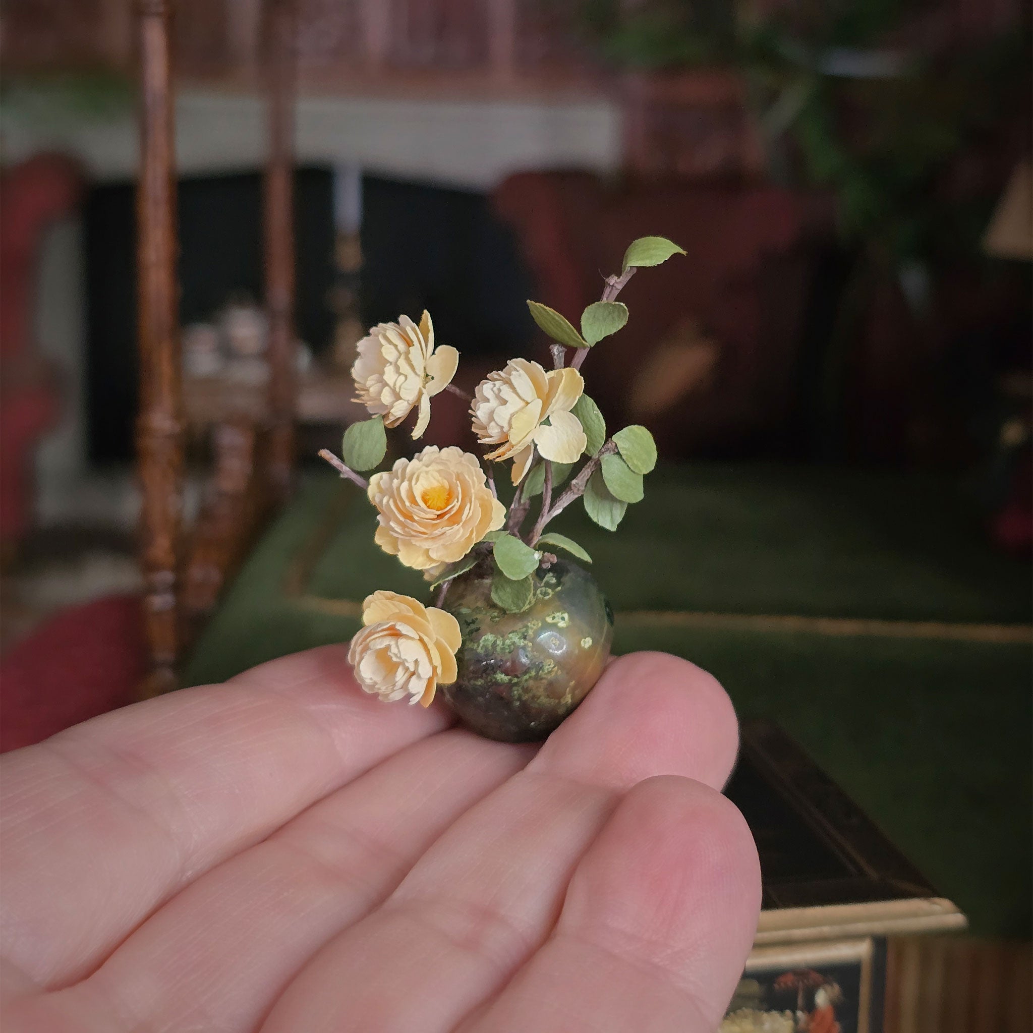 Hand holding a miniature vase with yellow flowers against a blurred indoor background