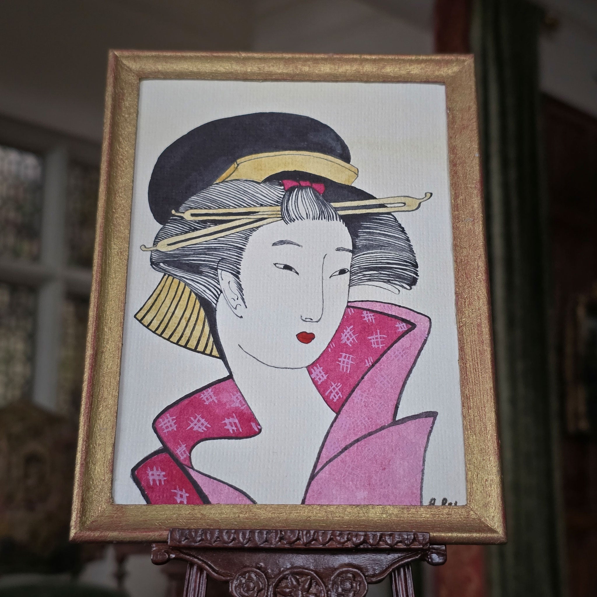 Framed artwork of a traditional Japanese geisha with a pink kimono and black hair, on a wooden easel. Alexandra R.