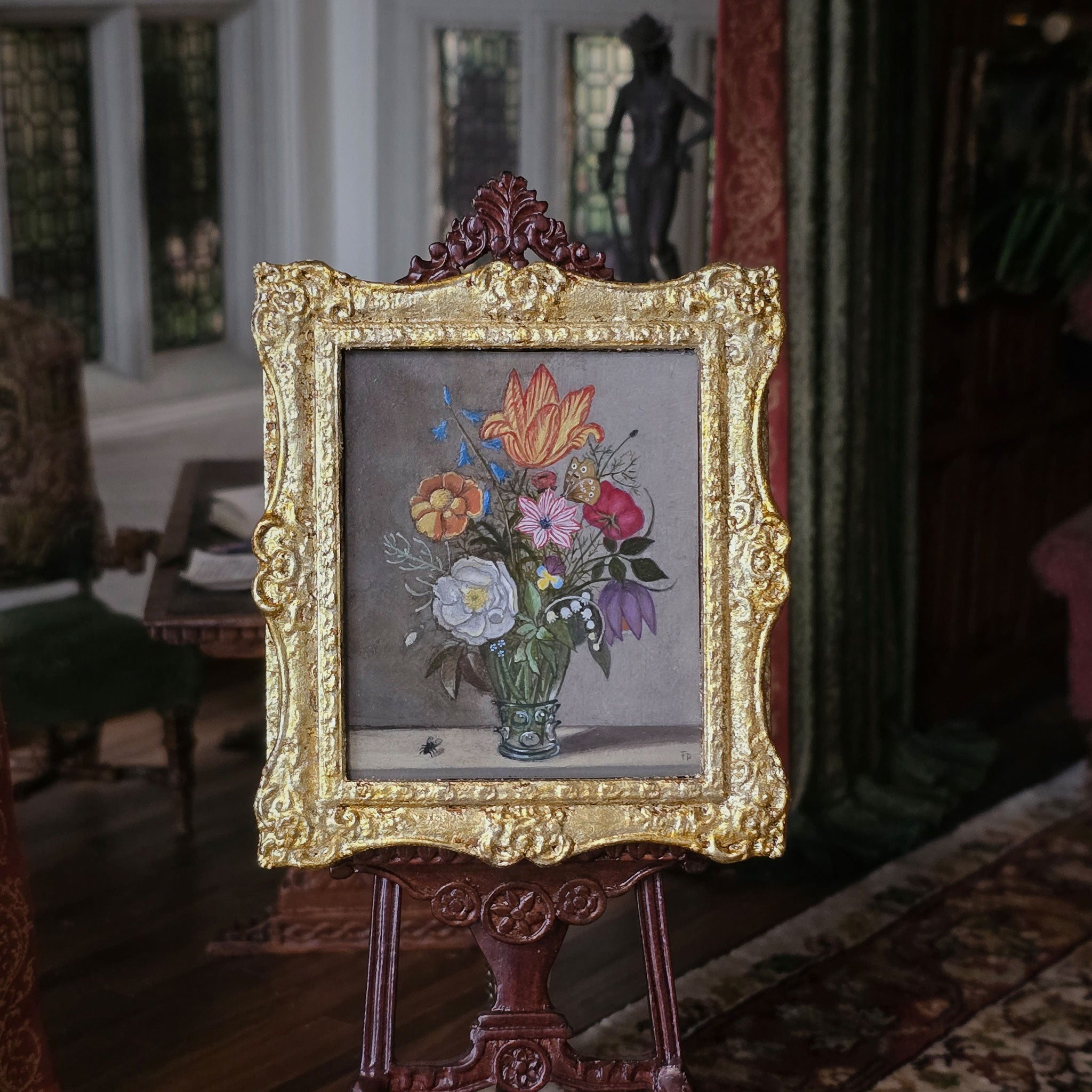 Decorative miniature painting still life of flowers in a gold frame on an easel in a room with classical decor.