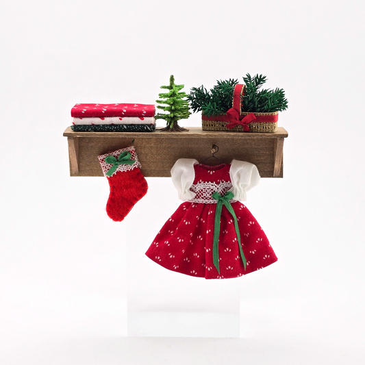 Dollhouse miniature  Red dress with white polka dots and green ribbon, red stocking, and small Christmas tree on a wooden shelf against a white background.