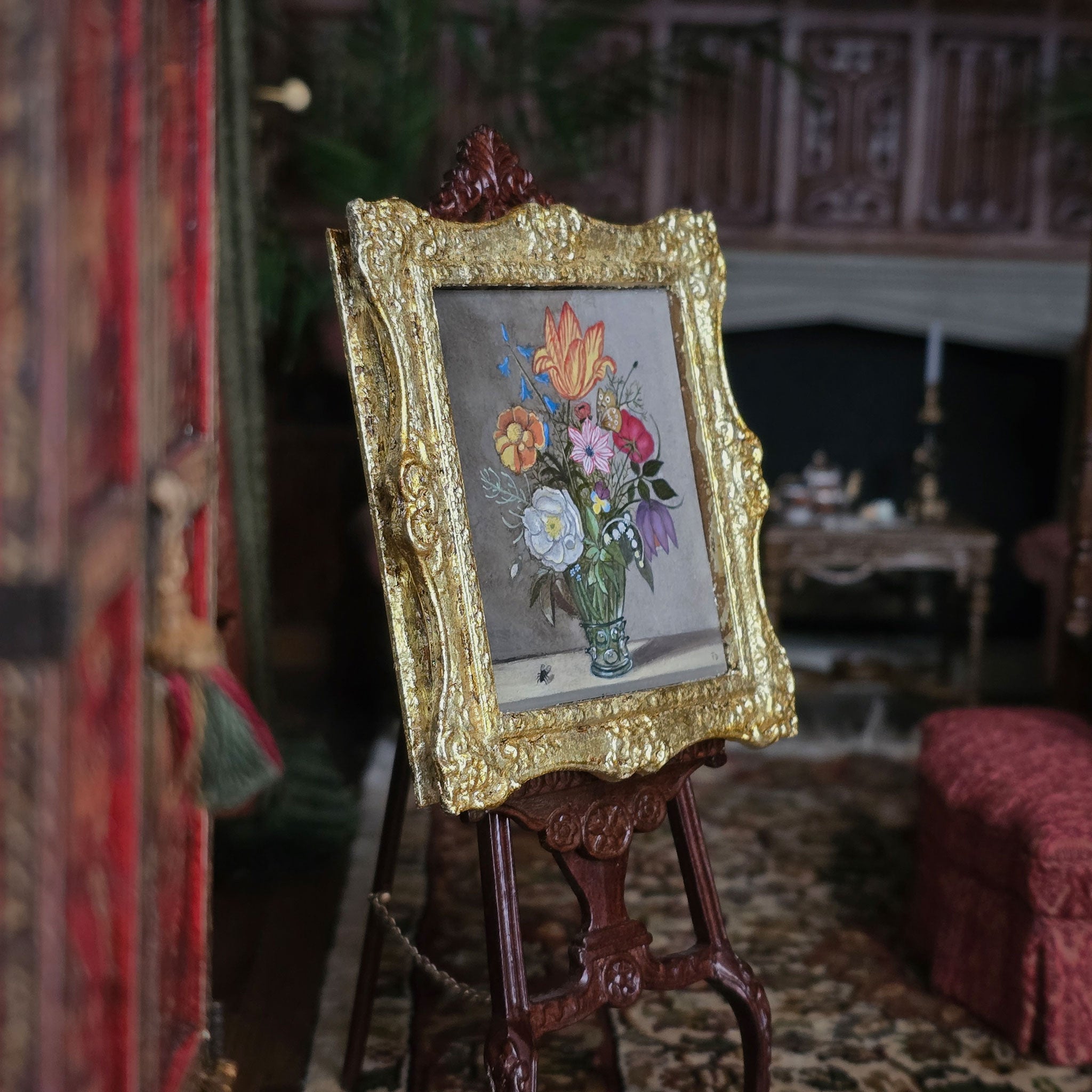 Miniature painting of flowers in a gold frame on an easel in a decorative room.
