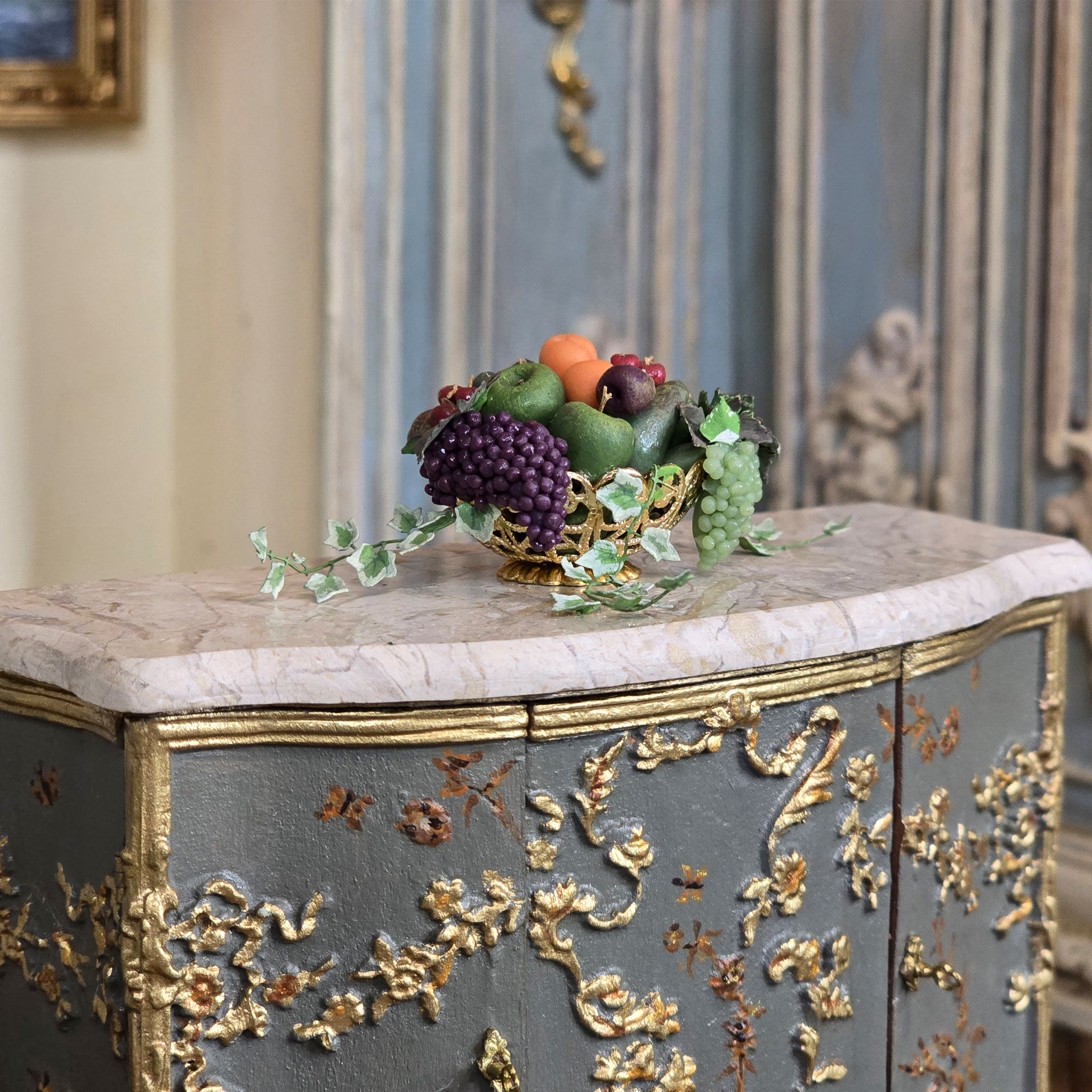 Decorative cabinet with gold leaf detailing and a bowl of fruit on a marble surface.