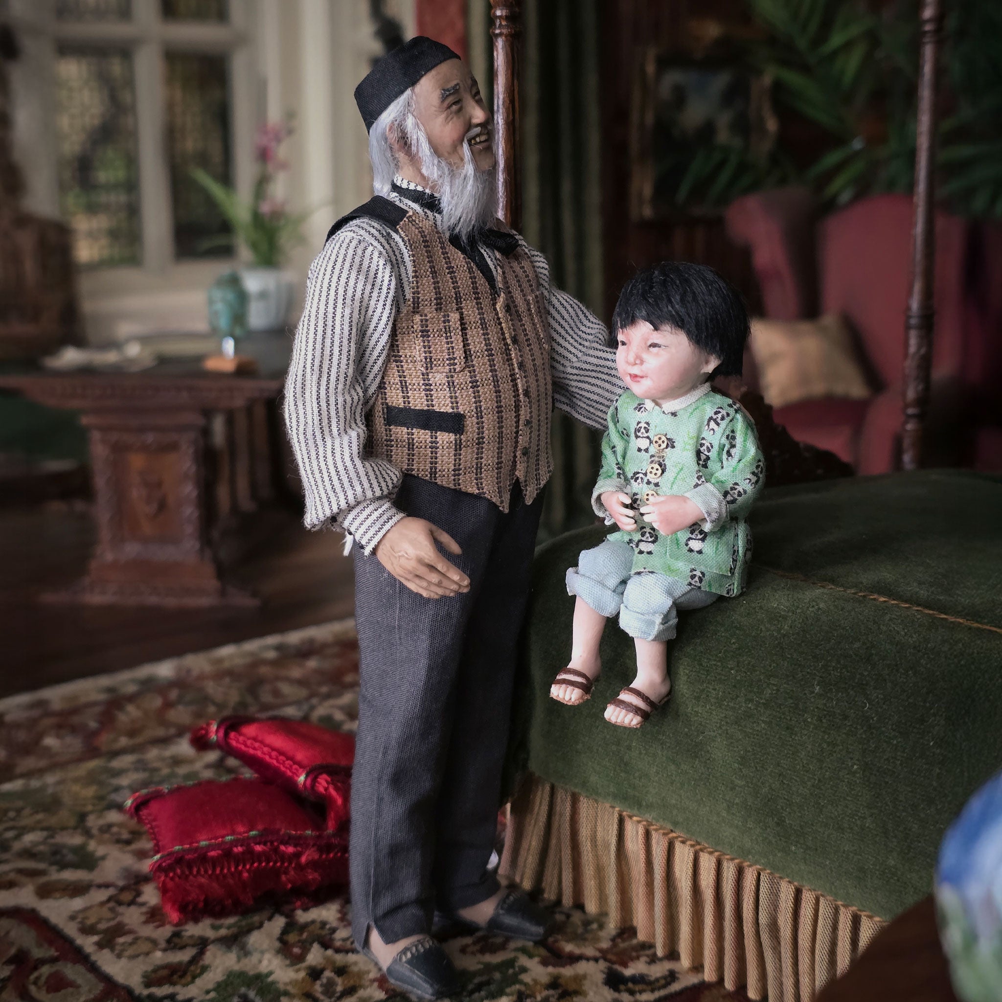 Two dolls, one Asian clay dollhouse doll and one grandchild, in a vintage-style room with furniture and decor.