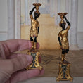 Load image into Gallery viewer, Two ornate pedestals with black figures, held by a hand in a dollhouse setting. John J. Hodgson
