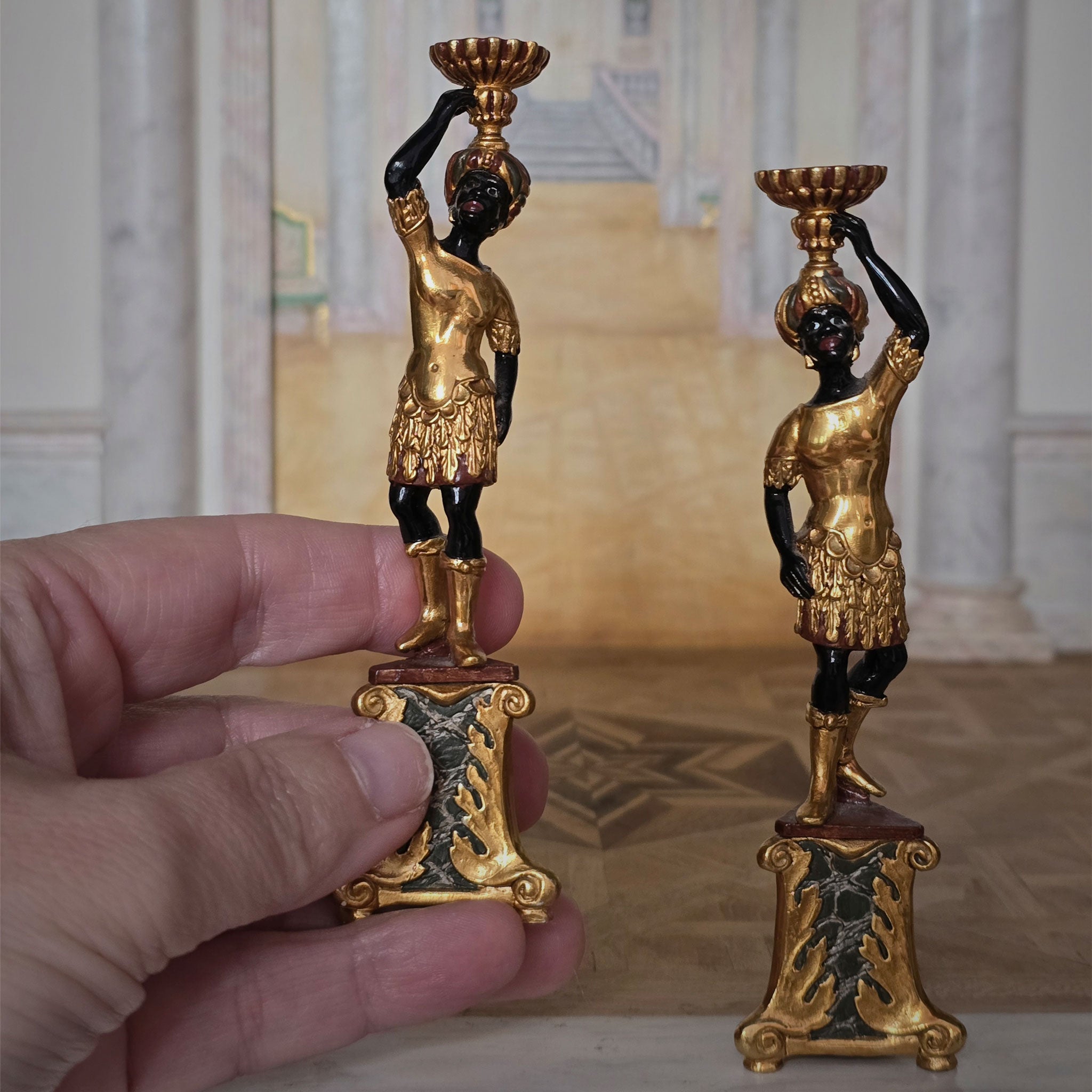 Two ornate pedestals with black figures, held by a hand in a dollhouse setting. John J. Hodgson